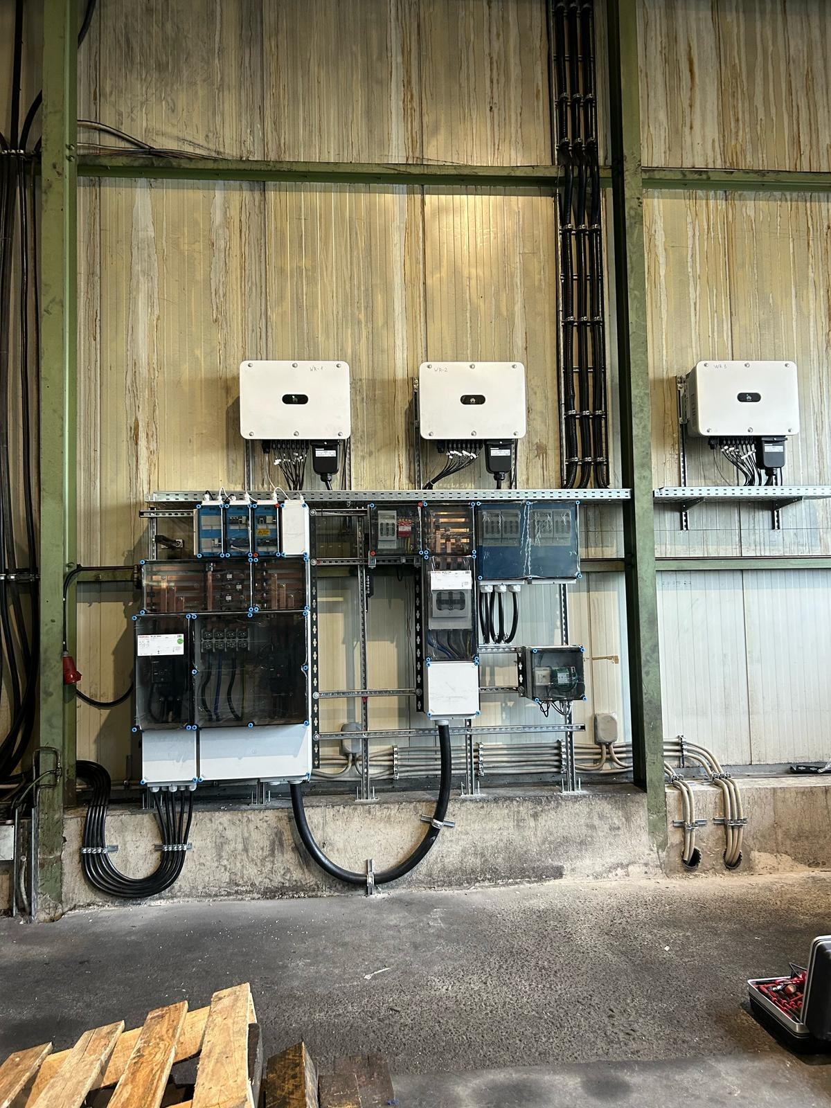 Zamontowane inwertery fotowoltaiczne i rozdzielnice na ścianie w hali przemysłowej, widoczne kable i przewody elektryczne.