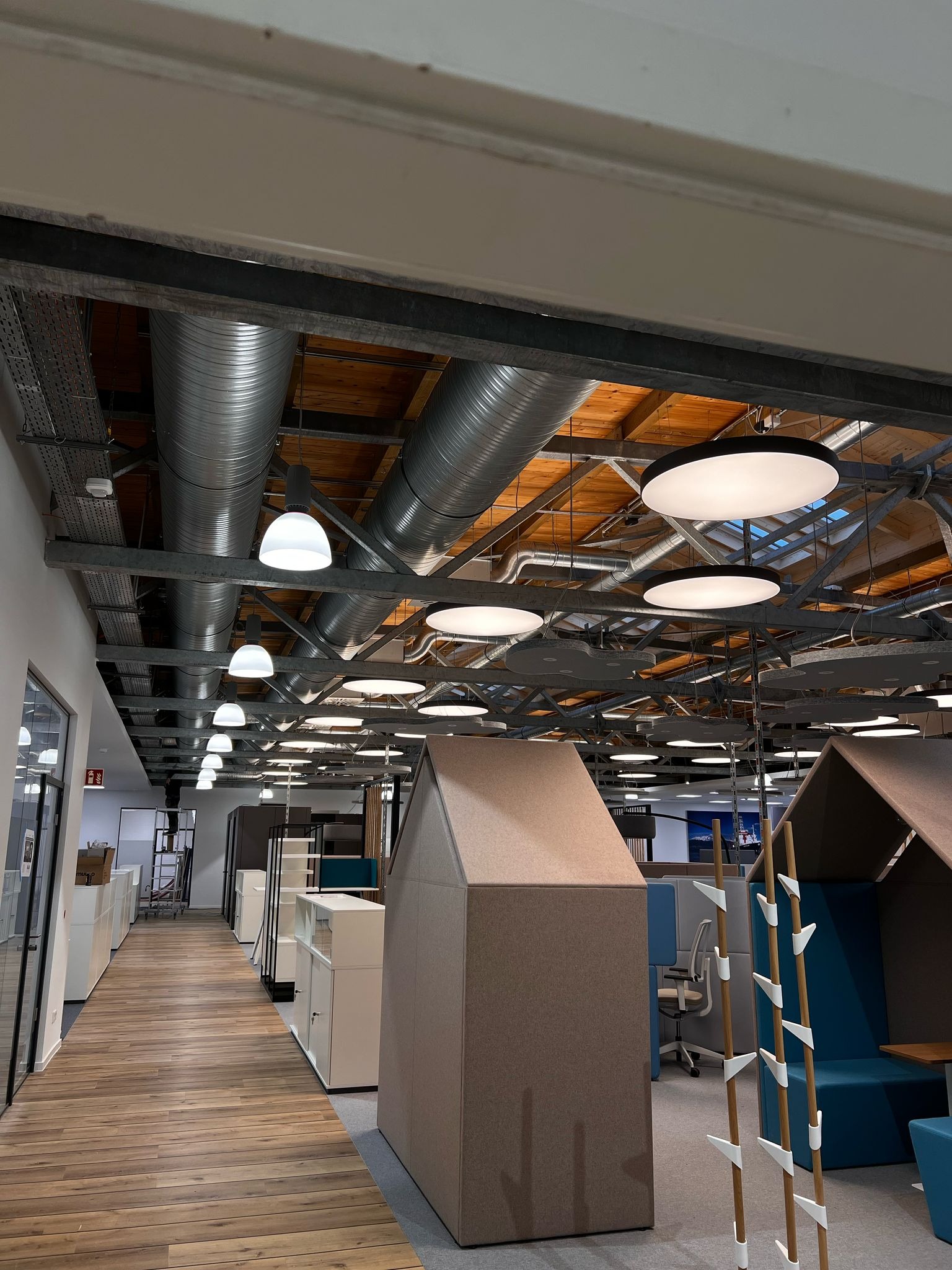 Oświetlenie biurowe: rzędy okrągłych lamp LED i rury wentylacyjne pod drewnianym sufitem w nowoczesnym biurze z kabinami akustycznymi i jasną podłogą.