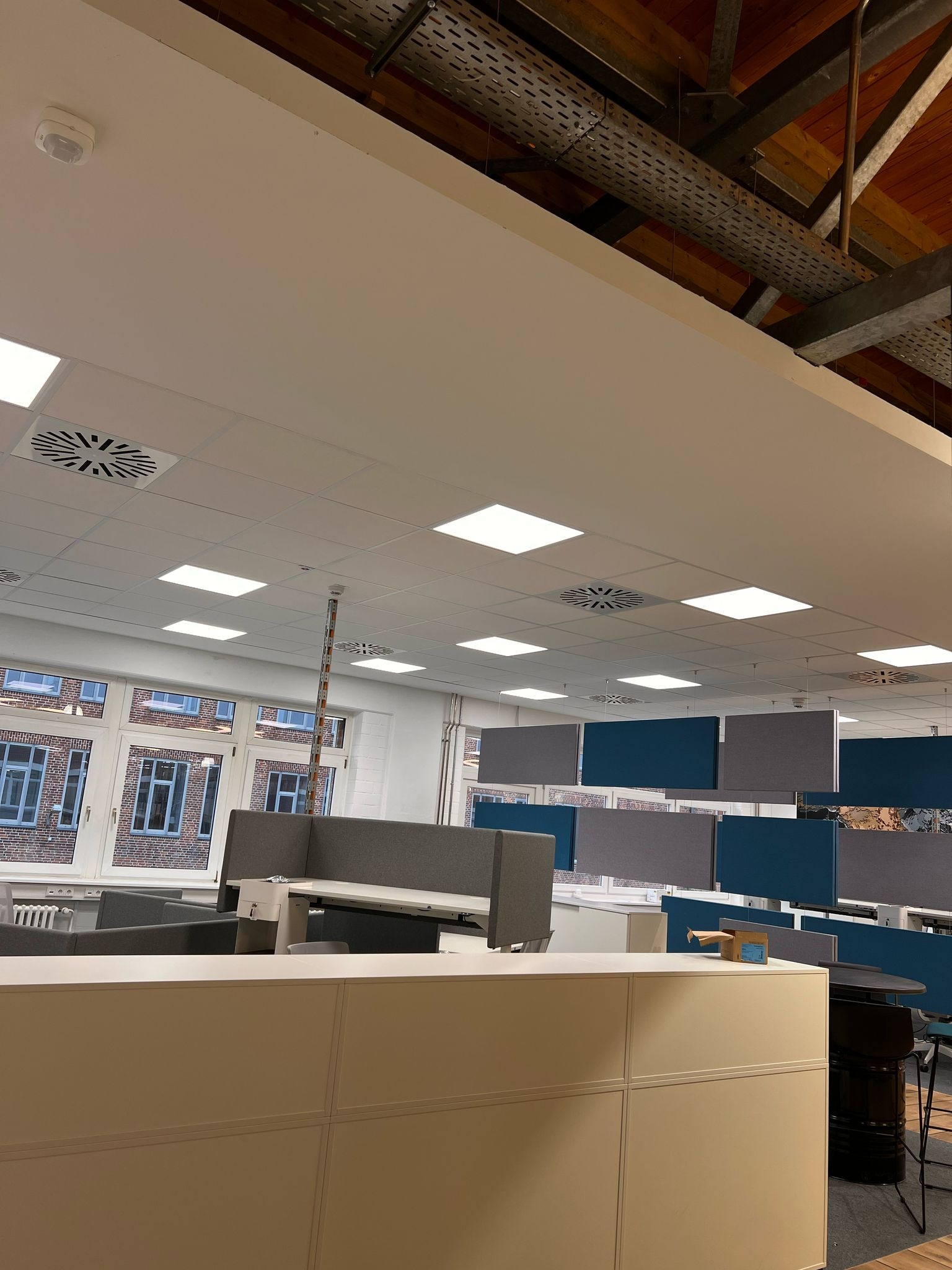 Nowoczesne biuro z otwartą przestrzenią, widoczne wentylatory sufitowe, instalacje pod sufitem i modułowe stanowiska pracy z panelami akustycznymi w odcieniach szarości i niebieskiego.