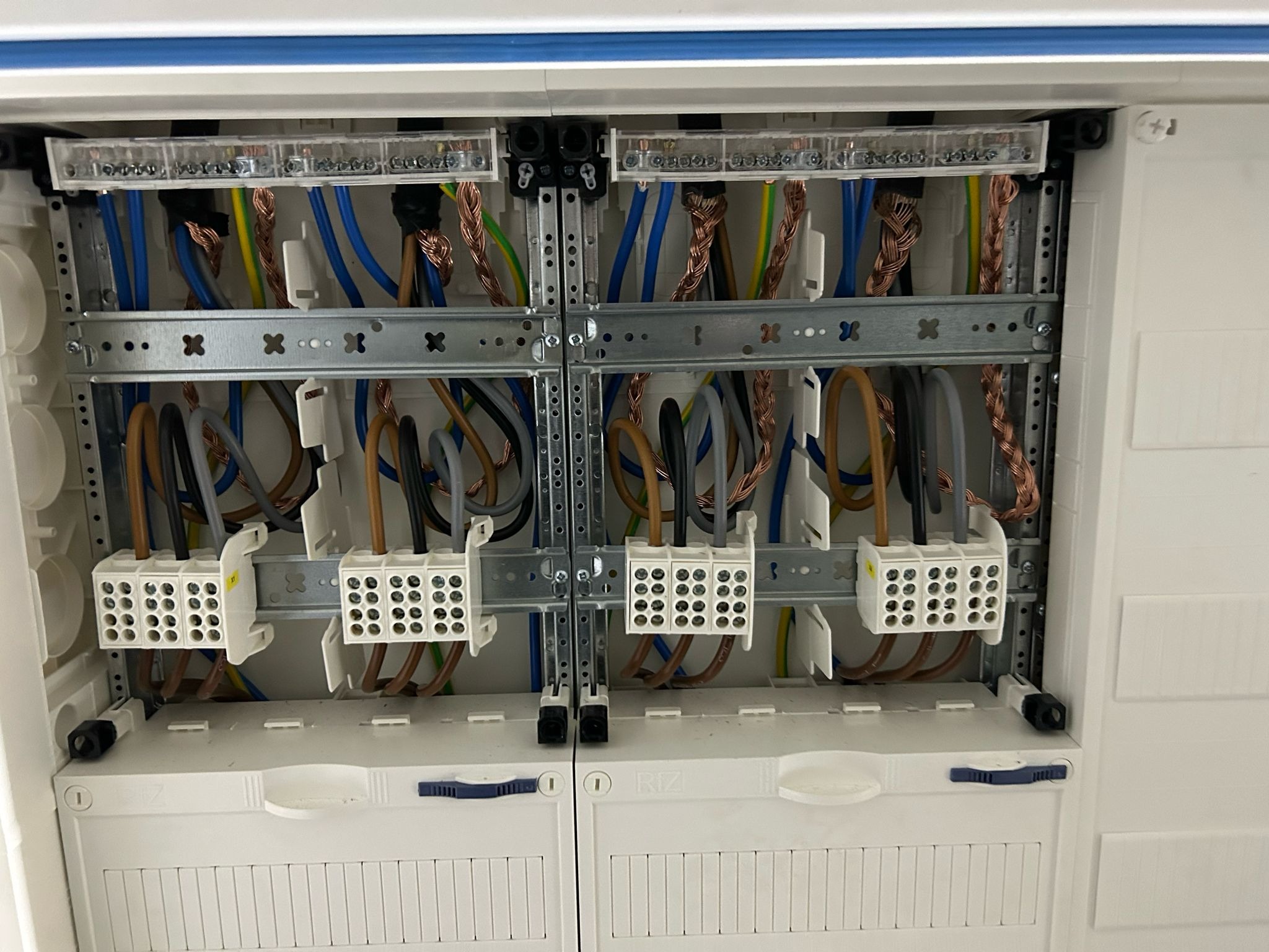 Rozdzielnica elektryczna z widocznymi kablami o różnych kolorach (niebieski, brązowy, czarny, zielono-żółty) podłączonymi do listew zaciskowych i szyn DIN w białej obudowie.