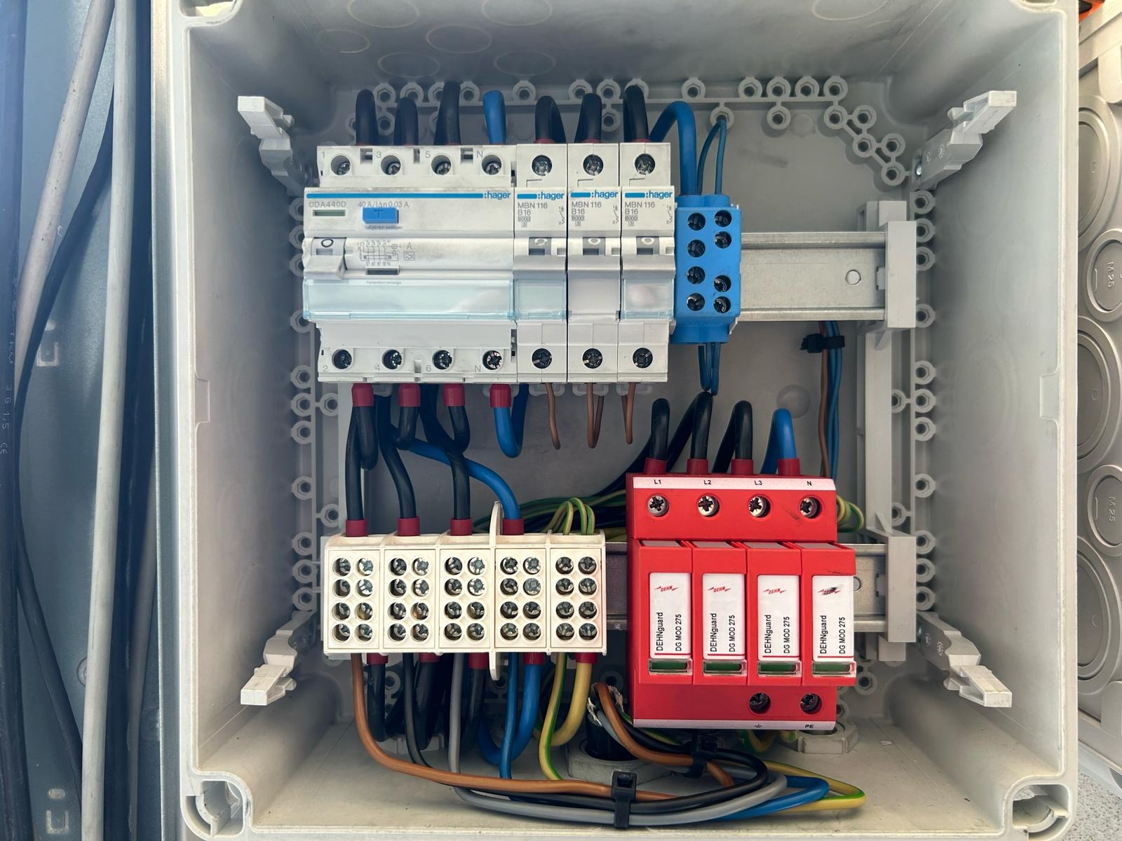 Rozdzielnica elektryczna w obudowie z widocznymi zabezpieczeniami przepięciowymi DehnGuard, bezpiecznikami Hager i listwami zaciskowymi, okablowanie w kolorach czarnym, niebieskim, brązowym...