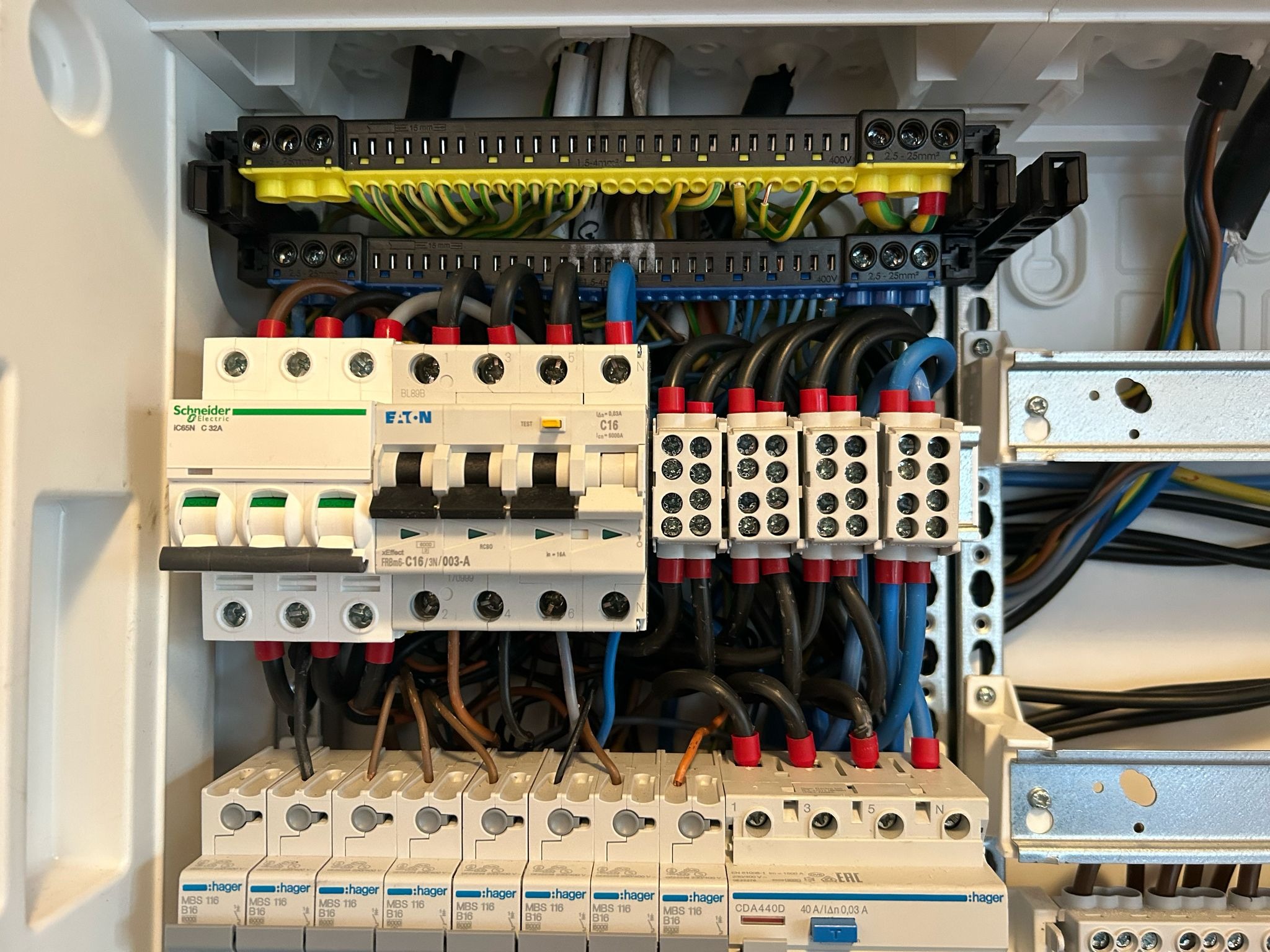 Rozdzielnica elektryczna z bezpiecznikami firm Schneider Electric i Eaton, widoczne kable w kolorach czarnym, niebieskim, brązowym i zielono-żółtym, ułożone wewnątrz białej obudowy.