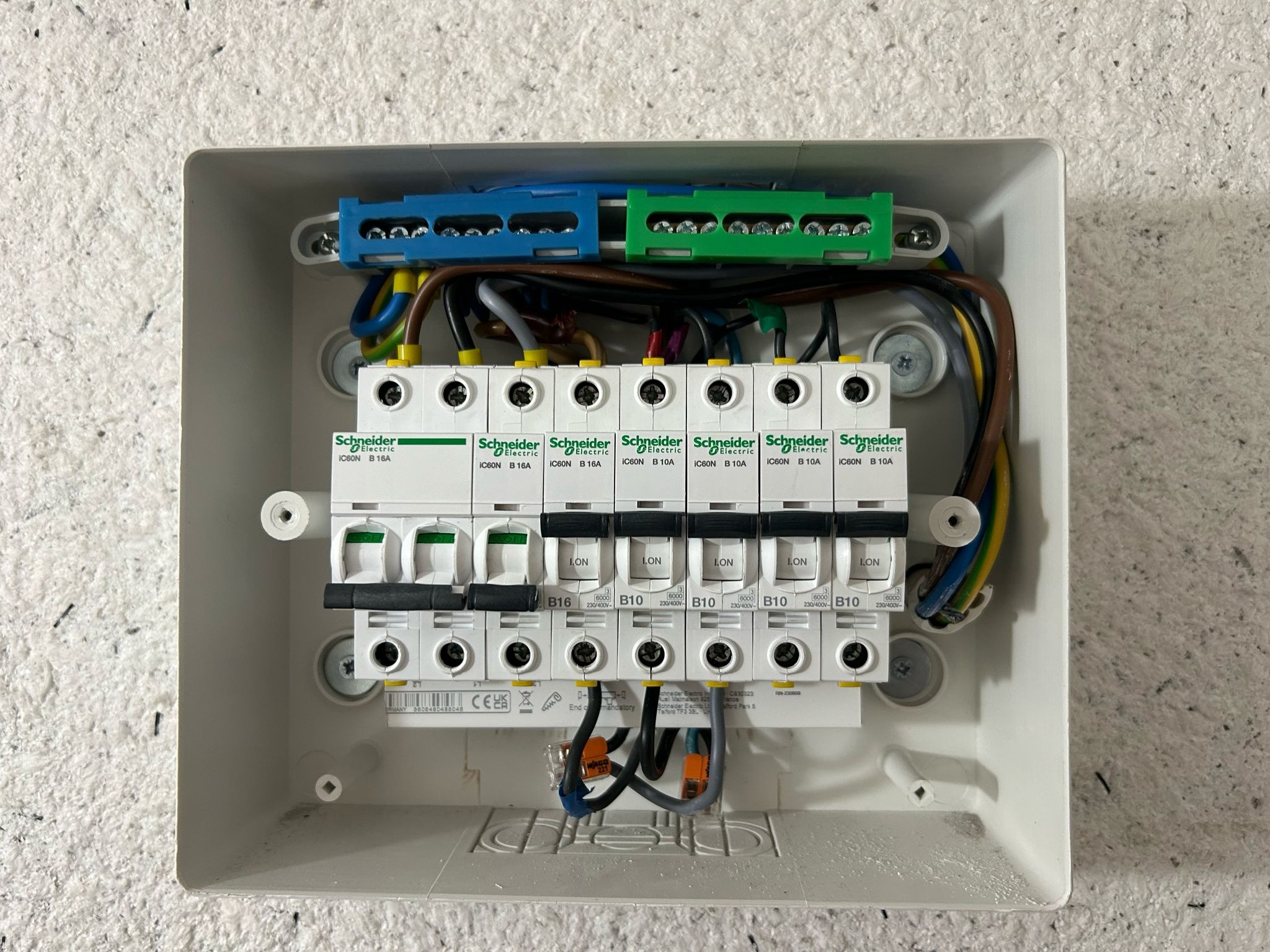 Rozdzielnica elektryczna z zainstalowanymi bezpiecznikami Schneider Electric IC60N B16 i B10, widoczne niebieskie i zielone listwy zaciskowe oraz przewody w izolacji brązowej, niebieskiej...