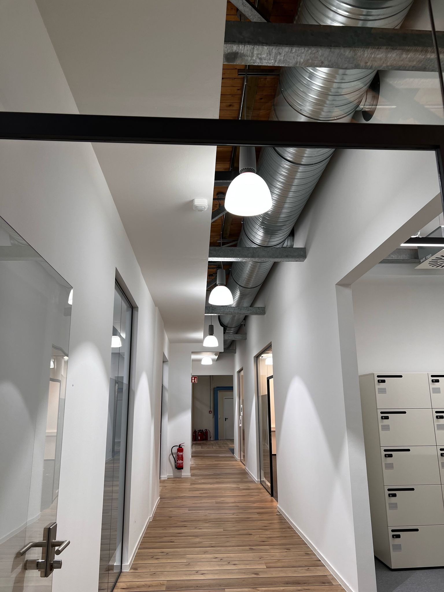 Długi korytarz biurowy z drewnianą podłogą, białymi ścianami, rzędem okrągłych lamp sufitowych i odsłoniętymi kanałami wentylacyjnymi pod sufitem.