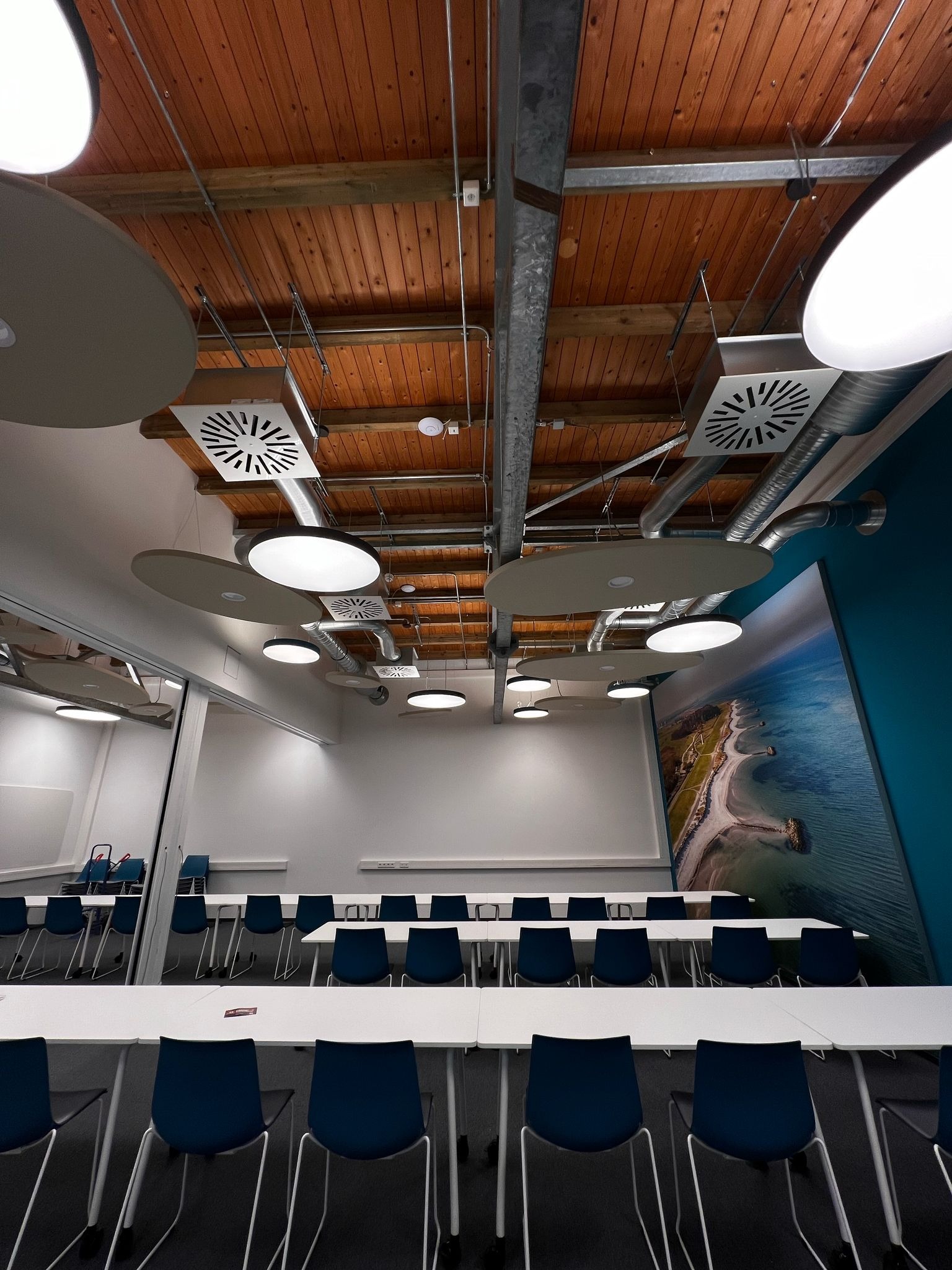 Sala konferencyjna z drewnianym stropem, okrągłymi lampami LED, systemem wentylacji i dużym obrazem plaży na ścianie.