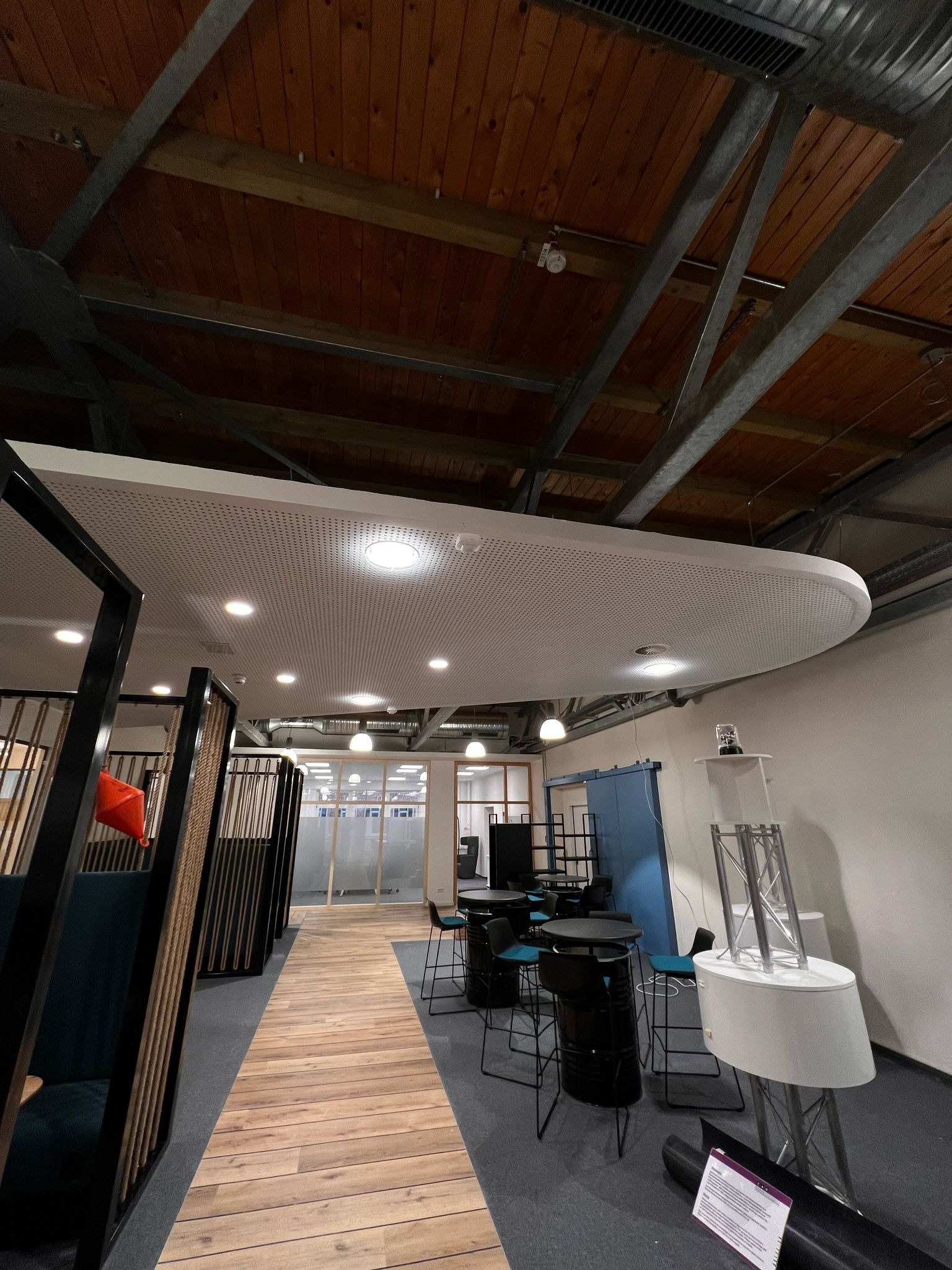 Nowoczesne biuro z drewnianą podłogą, metalową konstrukcją sufitu i oświetleniem punktowym, widok na przestrzeń otwartą z biurkami i krzesłami.