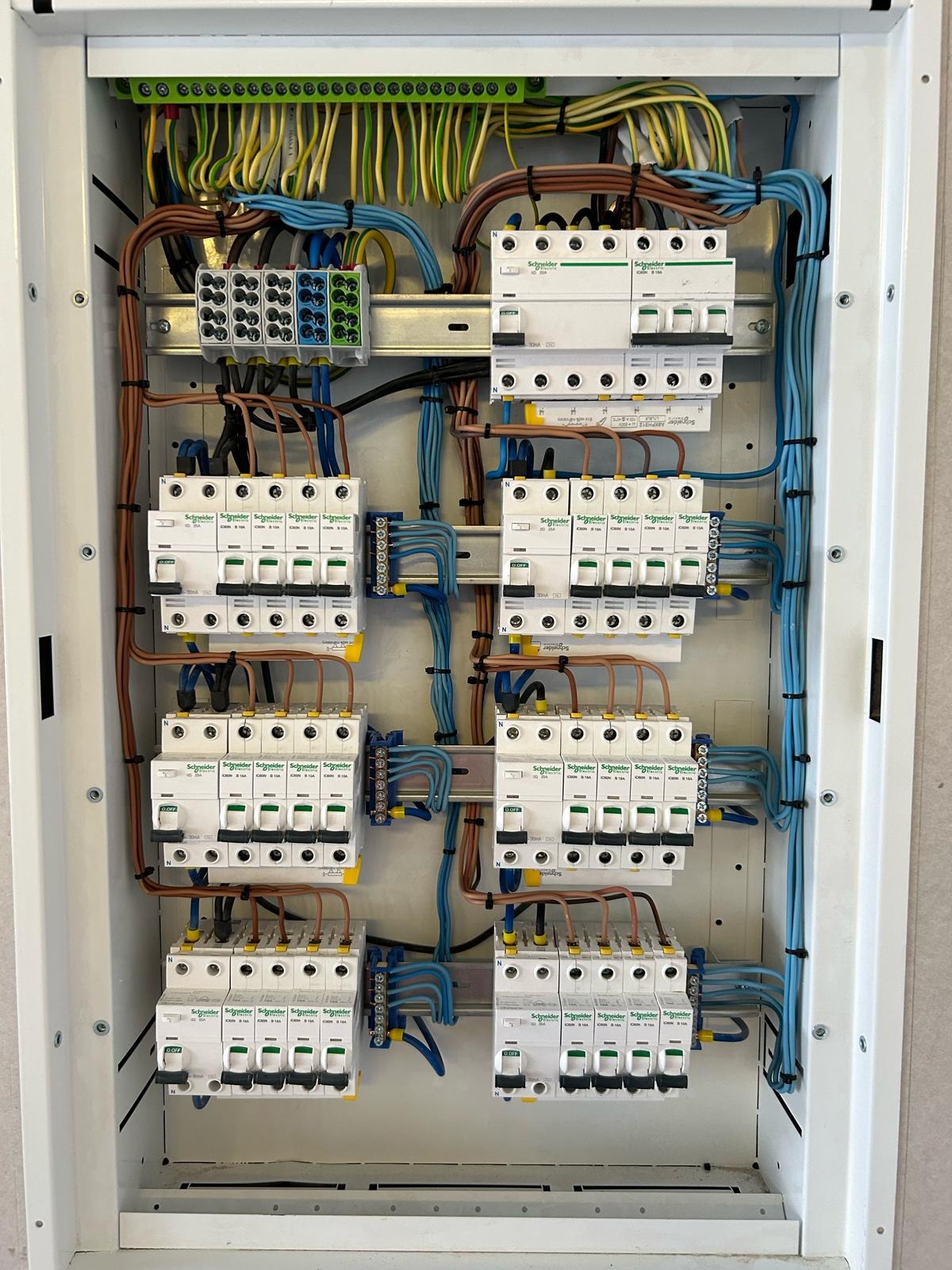 Rozdzielnica elektryczna z licznymi zabezpieczeniami Schneider Electric, starannie ułożone przewody w kolorach brązowym, niebieskim, żółto-zielonym, oraz listwy zaciskowe.