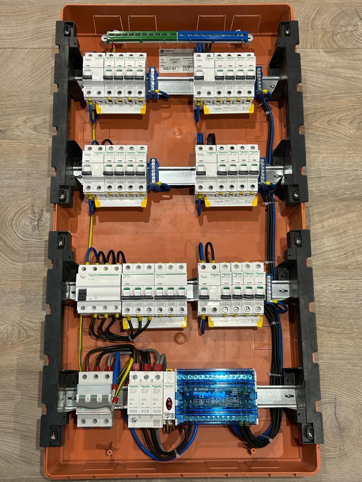 Rozdzielnica elektryczna z zamontowanymi bezpiecznikami i okablowaniem w pomarańczowej obudowie, widok z góry, różne modele i marki zabezpieczeń.