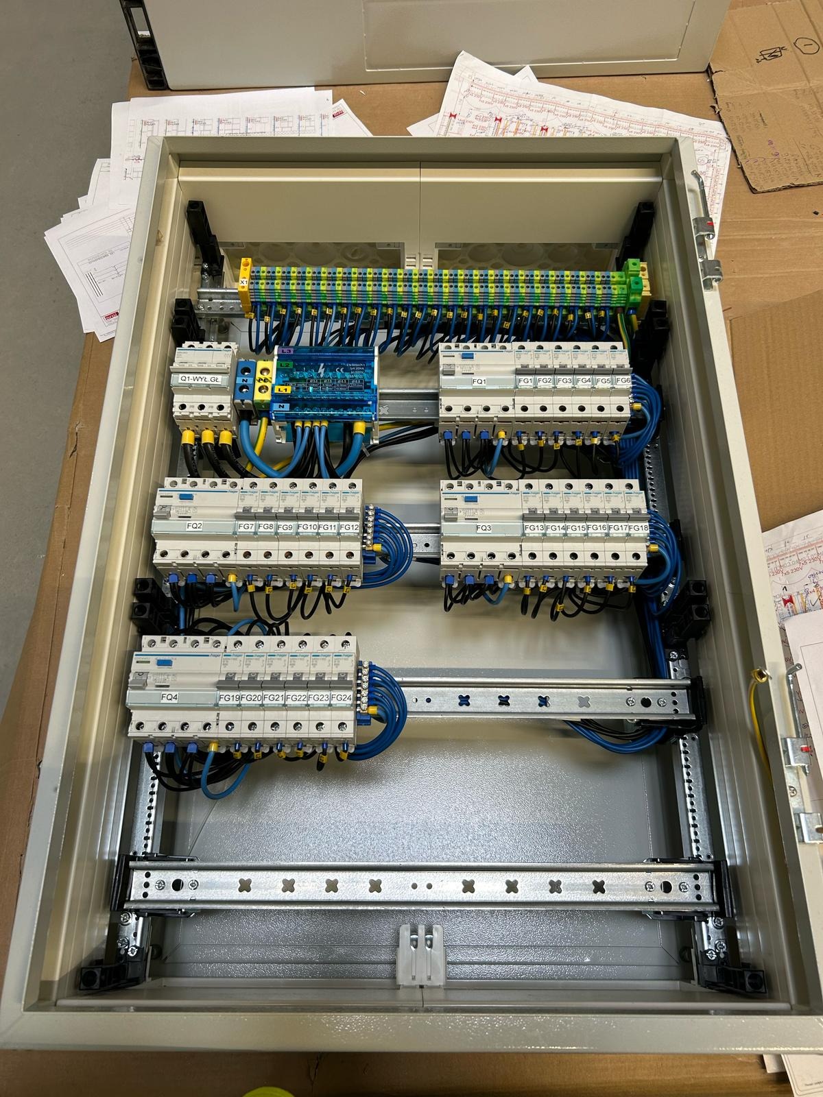 Otwarta szafa sterownicza z zamontowanymi bezpiecznikami, listwami zaciskowymi i okablowaniem w kolorach niebieskim, żółtym i czarnym. Widoczne opisy bezpieczników FQ1-FQ4 oraz FG1-FG24. Na górze...