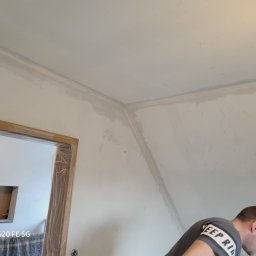 Kris-bau - Ściana w trakcie szpachlowania, widoczne ślady po nałożeniu gładzi, przygotowane do dalszej obróbki, zabezpieczona folią futryna drzwiowa, fragment sylwetki osoby w trakcie pracy.