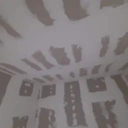 Surowy stan wnętrza z widocznymi łatami gipsowymi na ścianach i suficie, przygotowanymi do dalszych prac wykończeniowych, z przewodem zwisającym z sufitu.