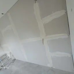 Ściana z płyt kartonowo-gipsowych, szpachlowane łączenia widoczne w zbliżeniu, aluminiowa drabinka oparta o ścianę, surowy beton na podłodze.