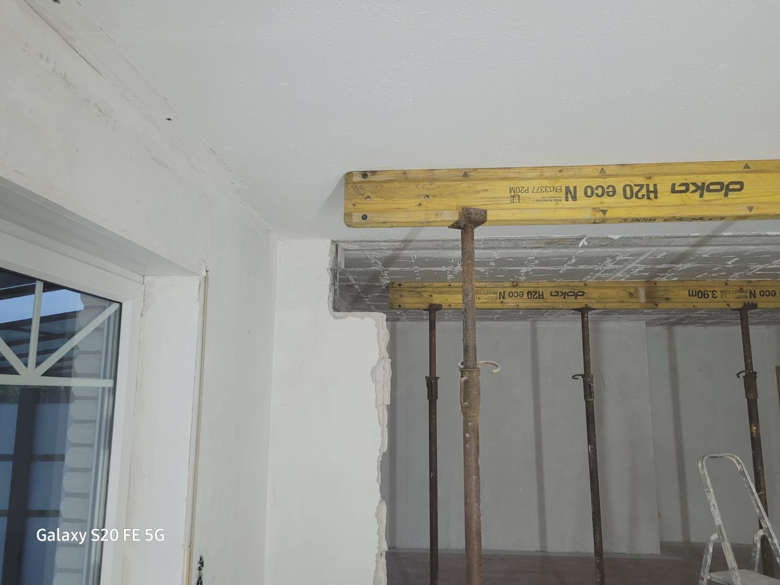 Wnętrze w trakcie prac przygotowawczych do wykończenia: widoczne stemple budowlane podtrzymujące strop, ściany z surowym tynkiem, okno z widokiem na zewnątrz.