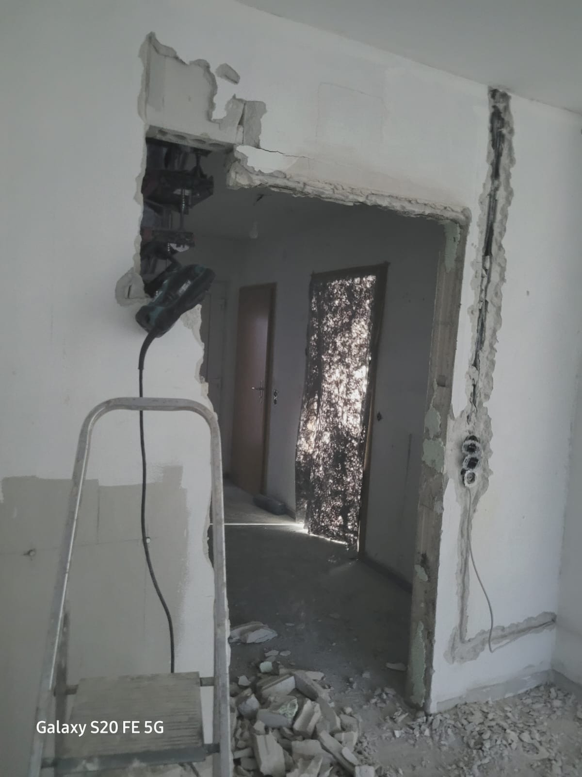 Demontaż ościeżnicy w trakcie remontu mieszkania, widoczny młot udarowy, fragment drabiny, gruz i odkryte przewody elektryczne w ścianie.