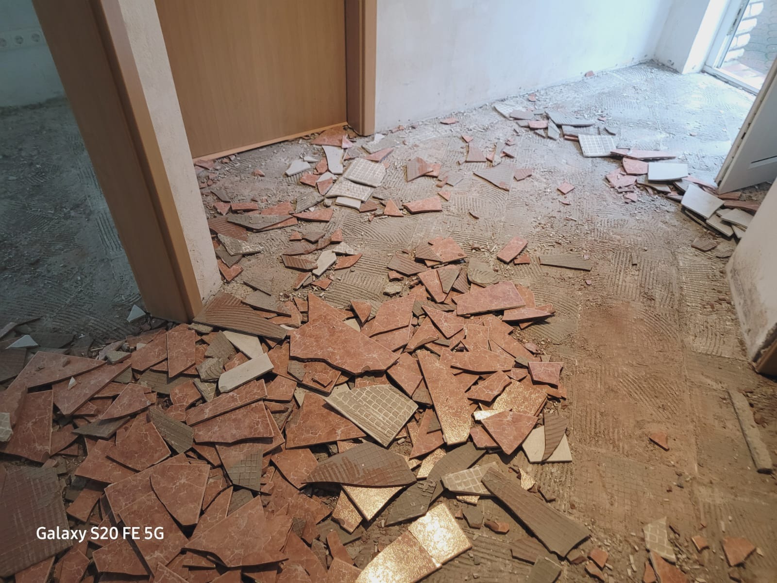 Fragment podłogi w trakcie demontażu starych płytek ceramicznych, widoczne odłamki w odcieniach czerwieni i brązu oraz ślady po kleju na podłożu, przy futrynie drzwi.