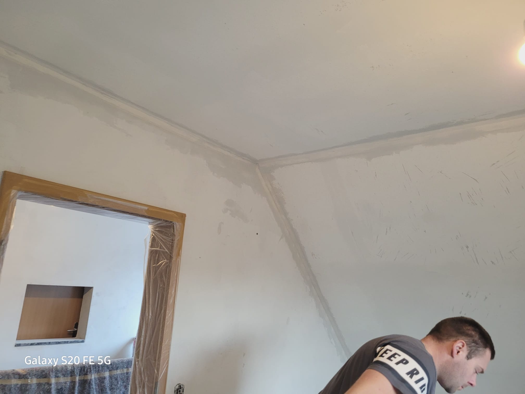 Ściana w trakcie szpachlowania, widoczne ślady po nałożeniu gładzi, przygotowane do dalszej obróbki, zabezpieczona folią futryna drzwiowa, fragment sylwetki osoby w trakcie pracy.