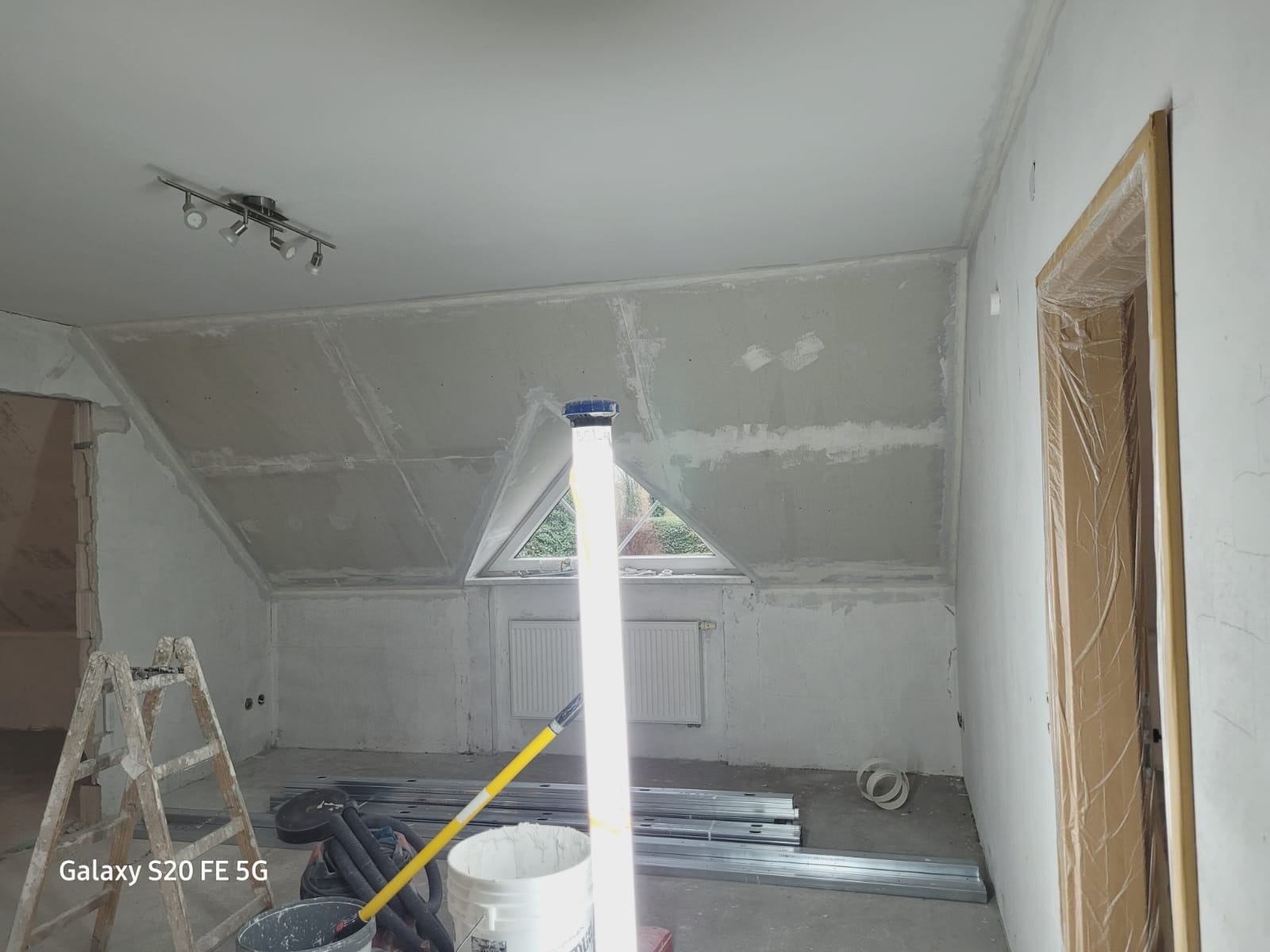 Poddasze w trakcie prac wykończeniowych: ściany z płyt kartonowo-gipsowych przed szpachlowaniem, widoczna drabina, profile metalowe, wiadro z farbą i lampa budowlana oświetlająca wnętrze. Okno...