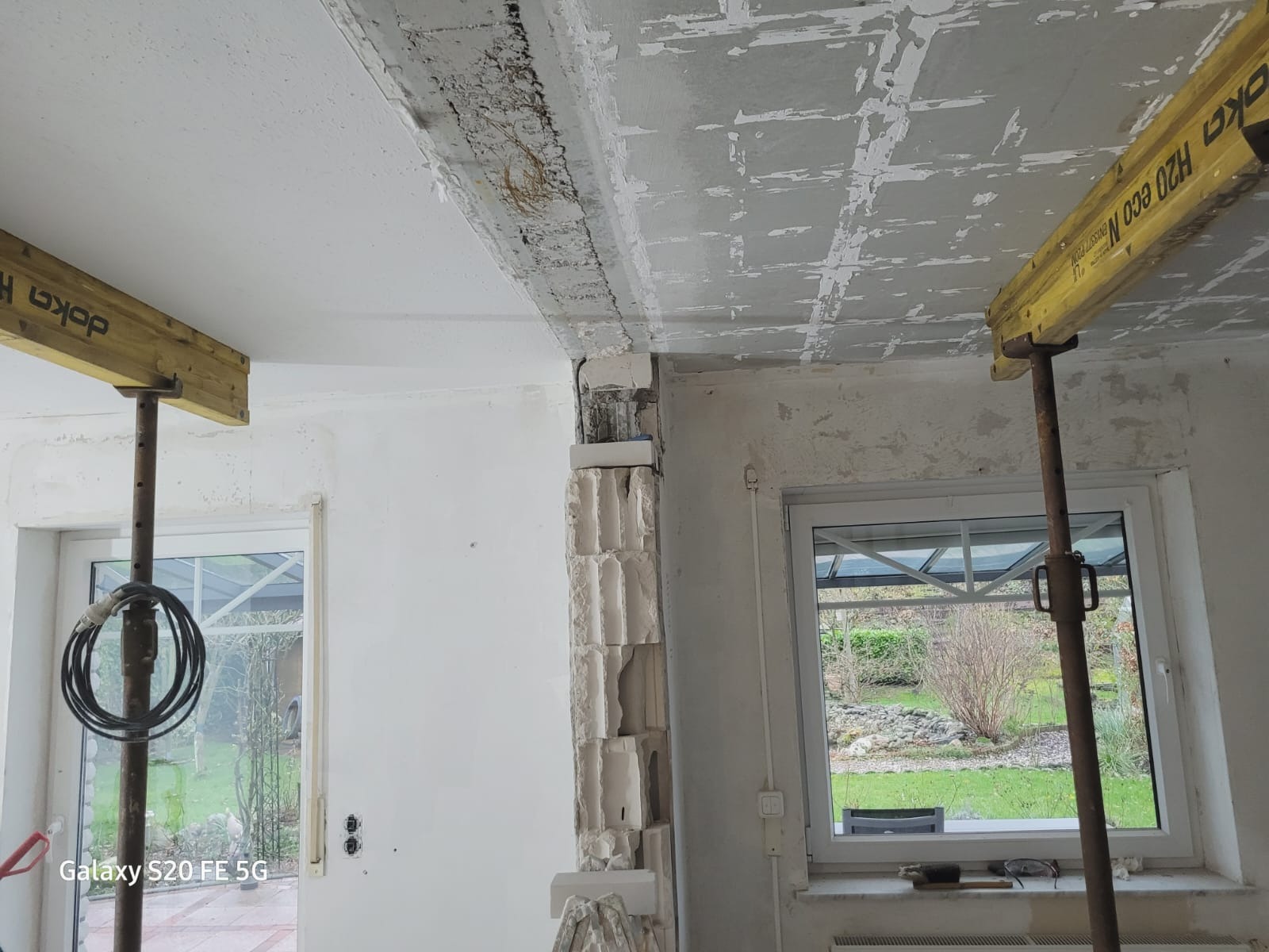 Wnętrze w trakcie prac remontowych: odsłonięta konstrukcja sufitu z widocznymi śladami po starej instalacji, podpora stropowa Doka H20 Eco N, fragment ściany z pustaków, okno z widokiem na ogród.