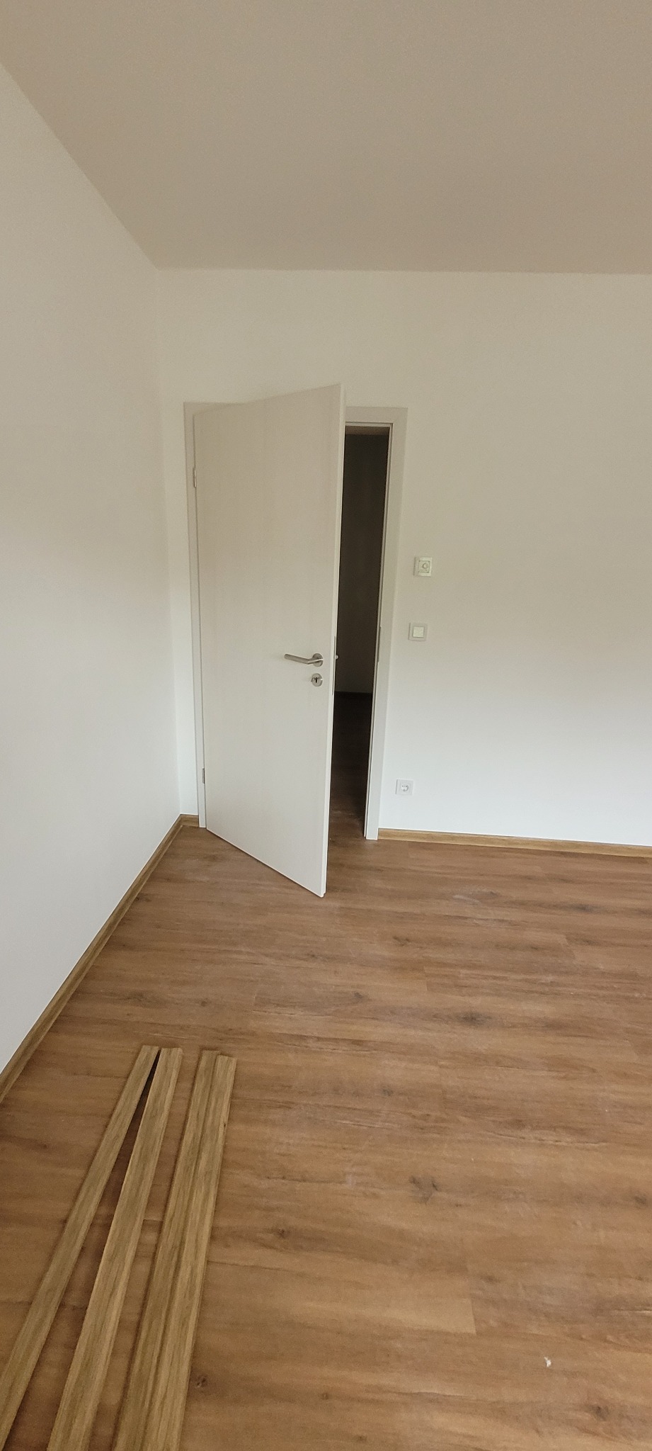 Jasne panele podłogowe w pokoju z białymi ścianami i otwartymi, białymi drzwiami. Widoczne listwy przypodłogowe. Naturalne, dzienne oświetlenie.