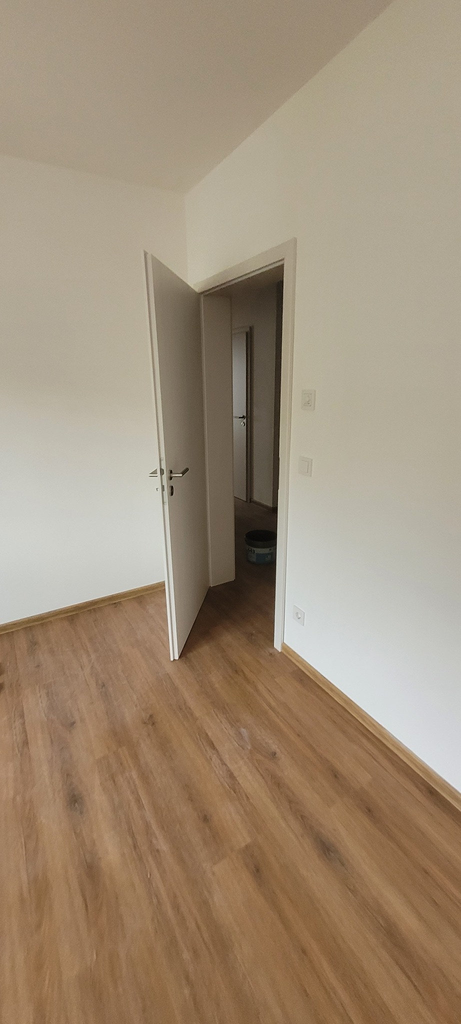 Jasne wnętrze po remoncie: świeżo ułożone panele podłogowe w odcieniach brązu, białe ściany i otwarte drzwi do kolejnego pomieszczenia. Widoczny fragment puszki z farbą.