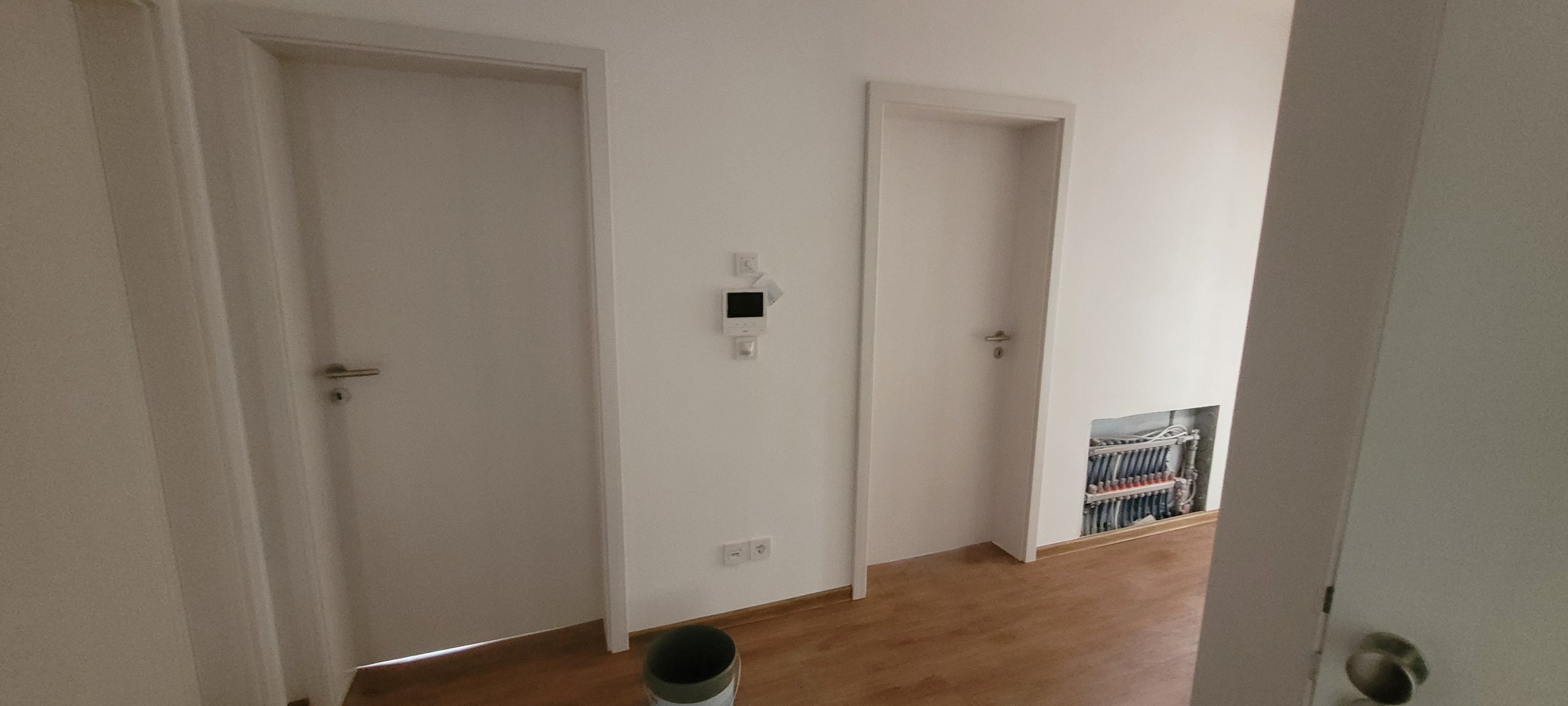 Jasne wnętrze po remoncie: dwie białe, zamontowane drzwi z srebrnymi klamkami, świeżo pomalowane ściany i fragment instalacji grzewczej w podłodze.