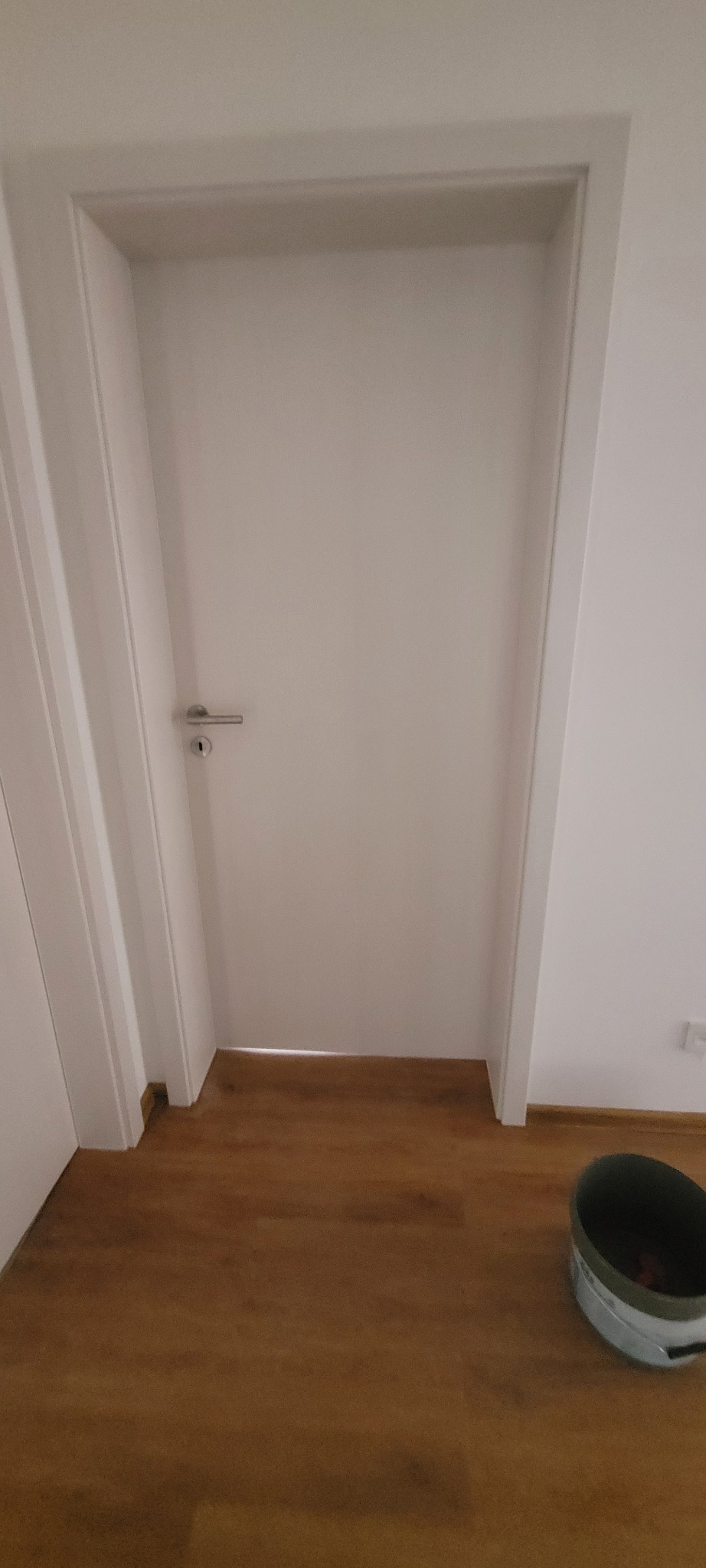 Białe drzwi wewnętrzne z srebrną klamką, widoczne panele podłogowe i wiadro z farbą w rogu. Minimalistyczny design, proste linie, świeżo po montażu.