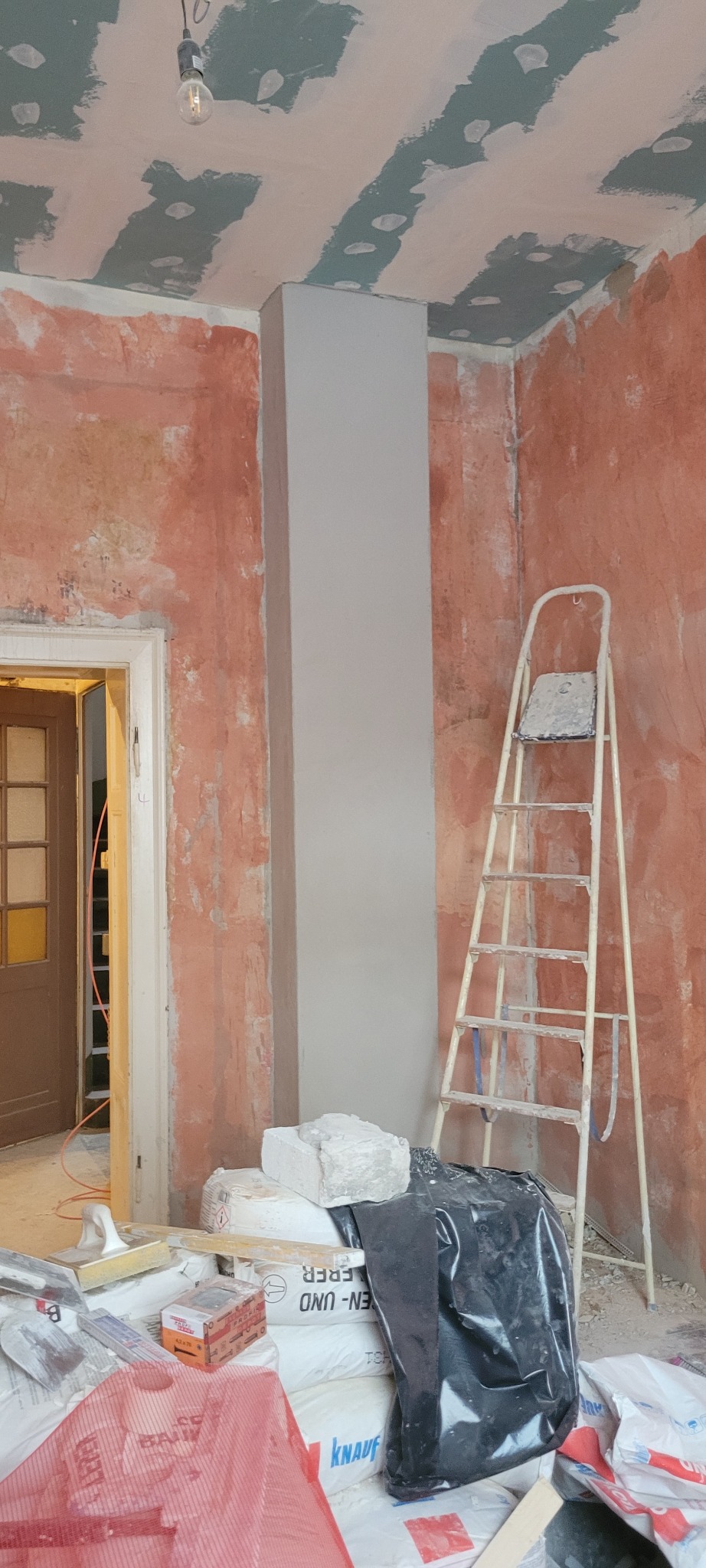 Pokój w trakcie remontu: ściany w surowym tynku, kolumna wyszpachlowana na gładko, drabina oparta o ścianę, worki gipsu i narzędzia na podłodze. Widoczne ślady prac szpachlarskich.