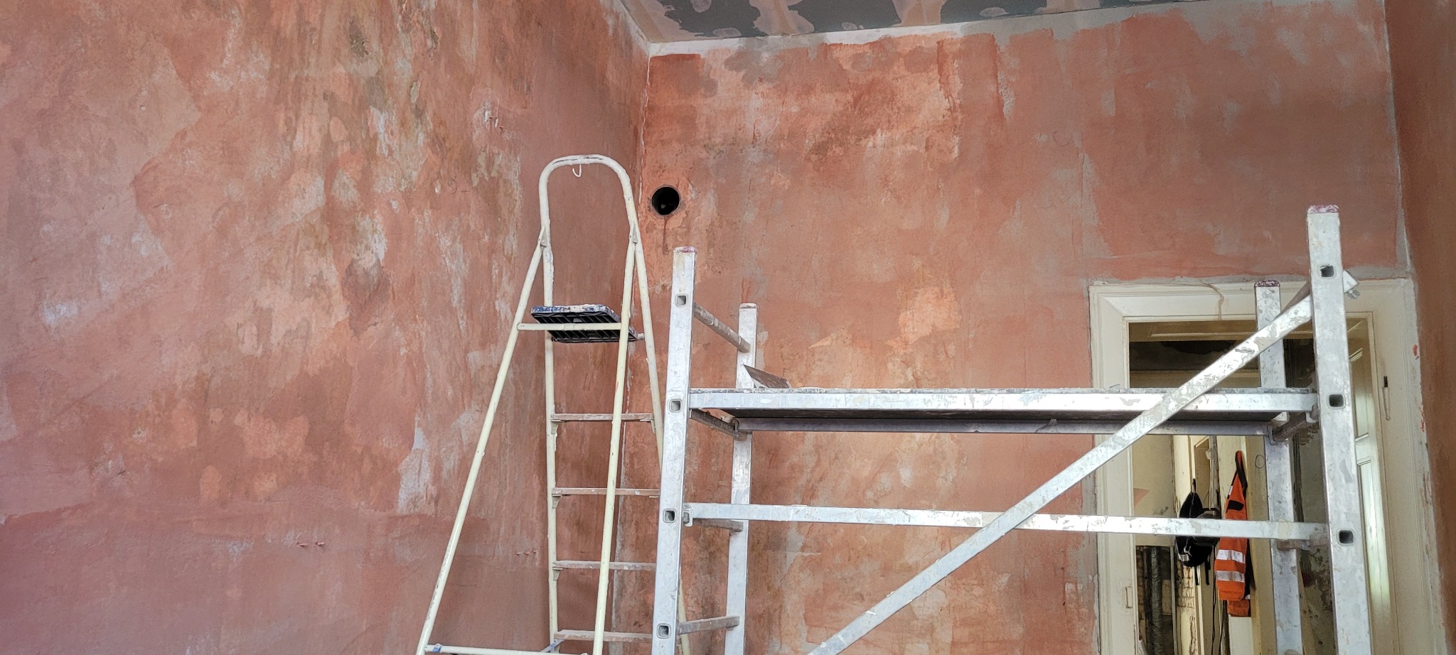 Ściana po szpachlowaniu w odcieniach różu, widoczne dwa rodzaje rusztowań: drabina i rusztowanie ramowe. Otwór w ścianie przygotowany pod instalację.