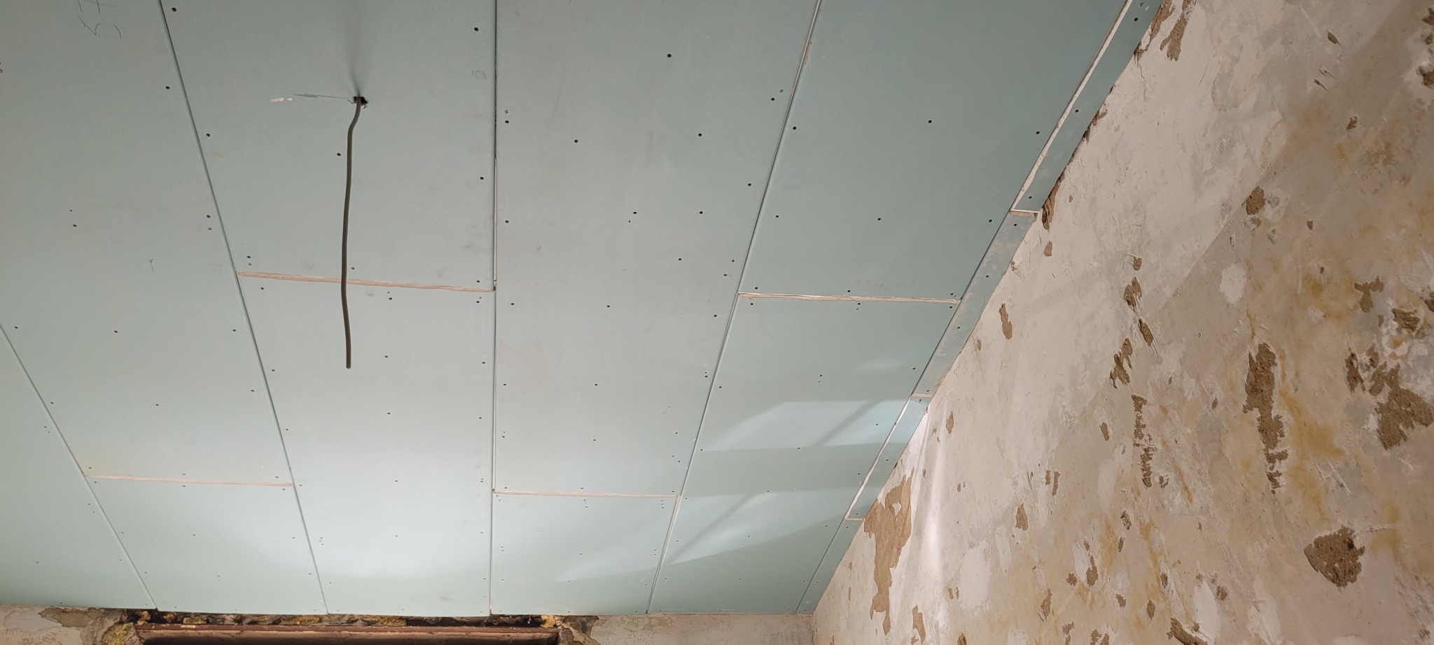 Montaż płyt gipsowo-kartonowych na suficie, widoczne łączenia i kabel elektryczny. Ściana w tle z widocznymi śladami po starym tynku. Prace wykończeniowe w trakcie.
