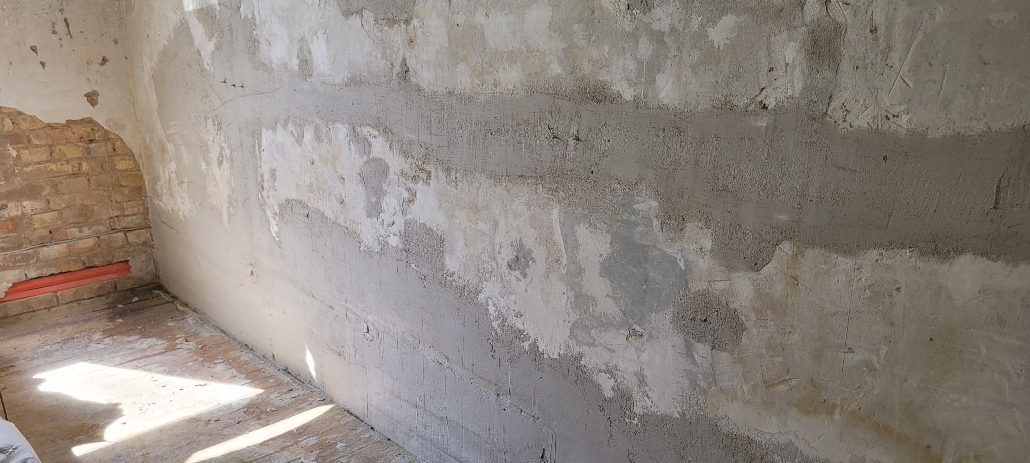 Ściana po szpachlowaniu z fragmentami cegły i instalacji. Widoczne nierówności i przygotowanie do dalszych prac wykończeniowych. Surowy stan wnętrza.