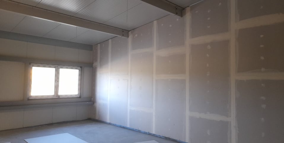 Ściany z płyt gipsowo-kartonowych, przygotowane do dalszych prac wykończeniowych. Widoczne łączenia płyt, szpachlowane i przeszlifowane. Okno w tle.