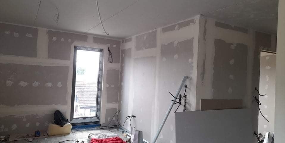 Wnętrze w trakcie szpachlowania: płyty gipsowe na ścianach, widoczne łączenia i otwór okienny. Na podłodze narzędzia i materiały budowlane.