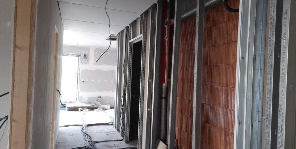 Wnętrze w trakcie prac wykończeniowych: metalowe profile na ścianach, instalacje, surowa cegła i przewody elektryczne. Widok na kolejne pomieszczenie.