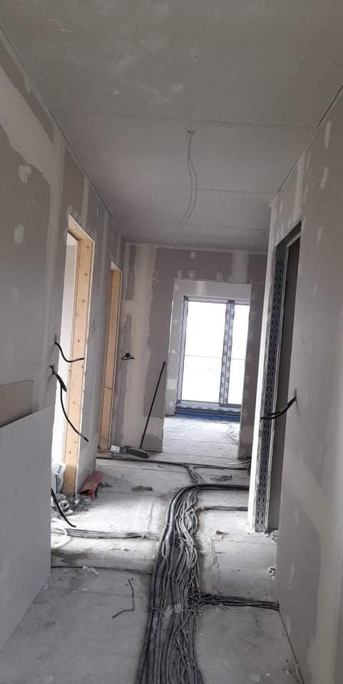 Wnętrze w trakcie szpachlowania i instalacji elektrycznej. Widoczne kable na podłodze, ściany przygotowane do malowania, ościeżnice drzwi i okno w tle. Surowy stan wykończenia.