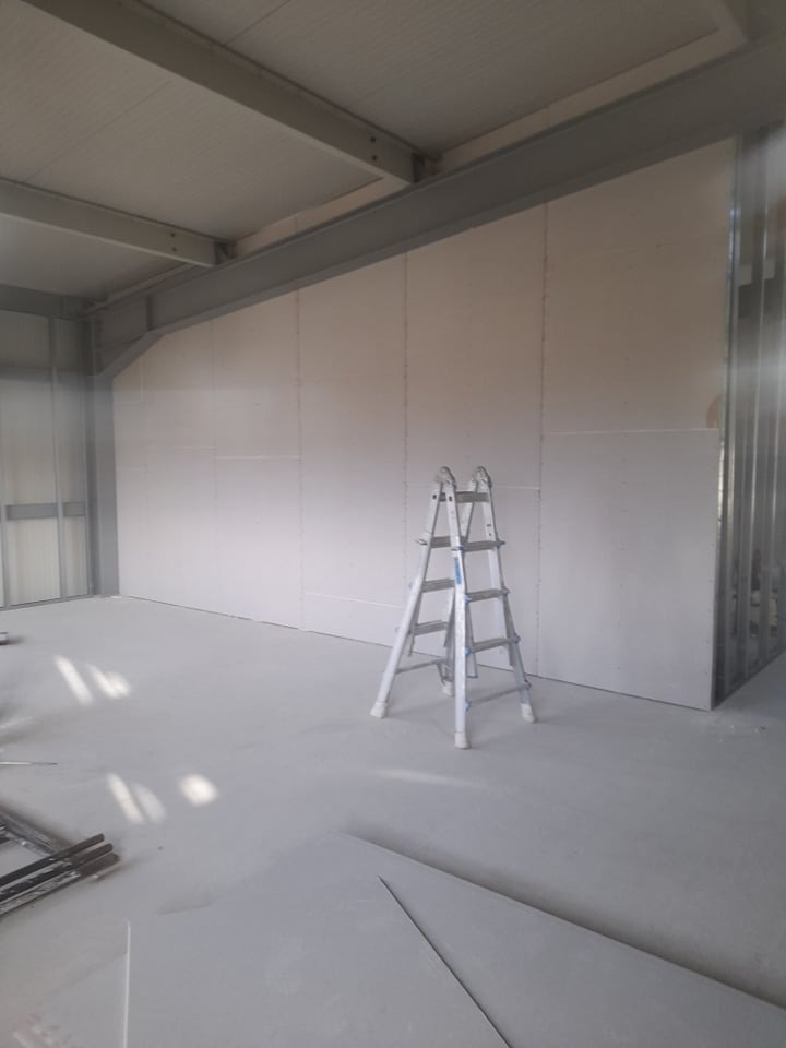 Wnętrze w trakcie szpachlowania: ściany z płyt gipsowych, drabina aluminiowa, metalowe belki stropowe. Widoczne płyty na podłodze. Surowy, industrialny charakter.