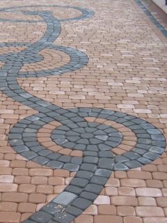 Ułożona nawierzchnia z kostki brukowej w odcieniach beżu i szarości, tworząca wzór wijącej się wstęgi z okrągłymi elementami. Kostka ułożona regularnie, widoczne spoiny. Perspektywa z dołu do góry.