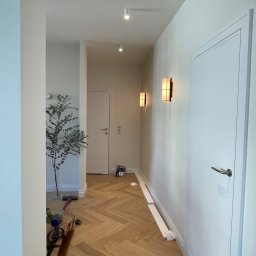 INVEST BUD Marcin Pyc - Wnętrze mieszkania w trakcie prac wykończeniowych: widoczna podłoga ułożona w jodełkę, białe ściany z zamontowanymi lampami ściennymi, drzwi wewnętrzne, narzędzia i materiały budowlane na podłodze,...