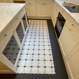 INVEST BUD Marcin Pyc - Wykończona kuchnia z kremowymi szafkami, marmurowym blatem i wzorzystą podłogą z płytek w odcieniach szarości i bieli, kontrastującą z ciemną, gumową podłogą graniczącą z parkietem.