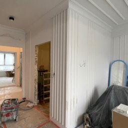 INVEST BUD Marcin Pyc - Wnętrze w trakcie prac wykończeniowych: ściany z dekoracyjnymi listwami pionowymi, widoczny odkurzacz przemysłowy, zabezpieczona podłoga, drabina w przejściu do innego pomieszczenia, elementy...