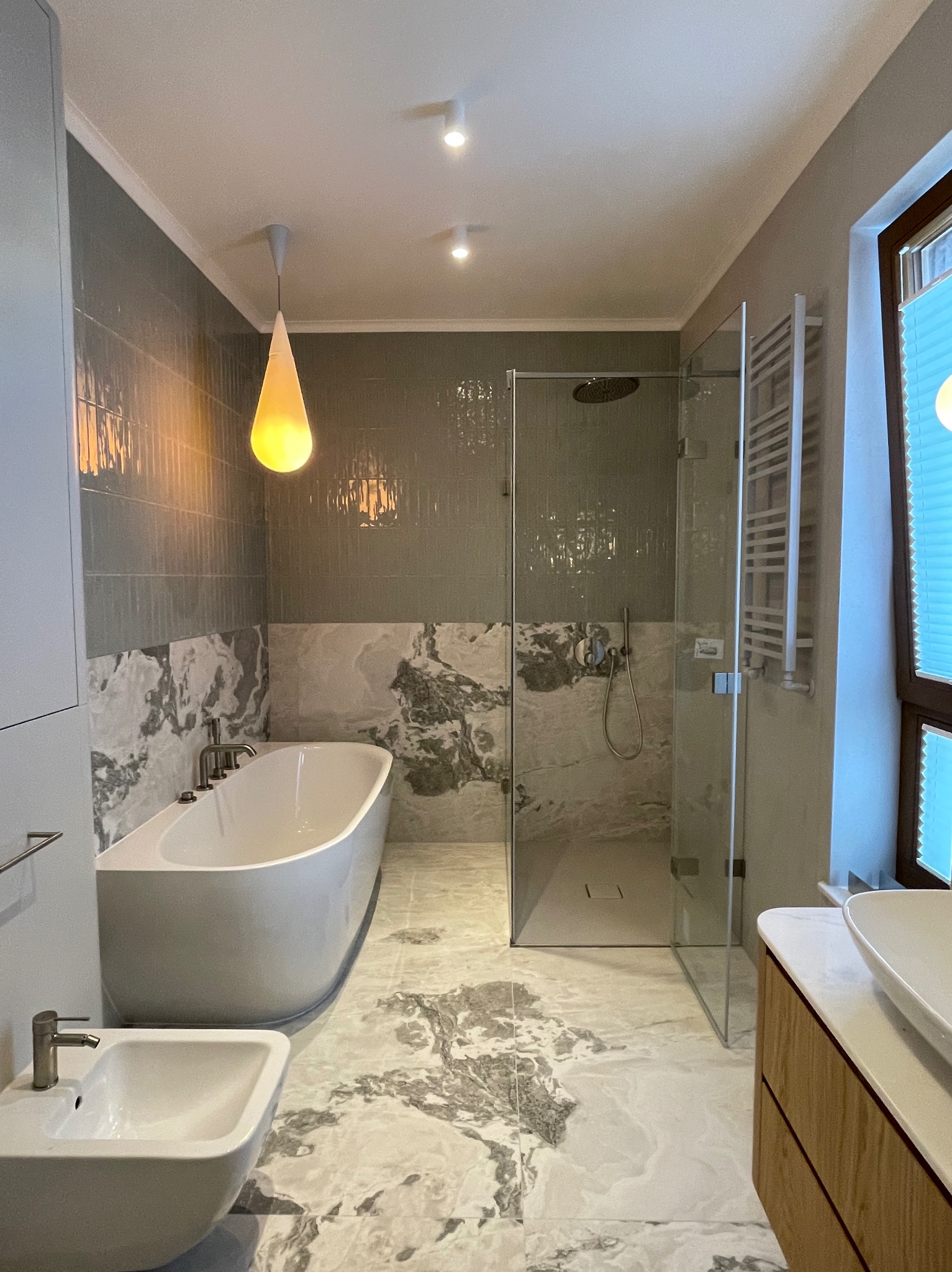 Nowoczesna łazienka z szarymi płytkami imitującymi cegłę, marmurową podłogą i wolnostojącą wanną, oświetlona wiszącą lampą w kształcie kropli i halogenami.