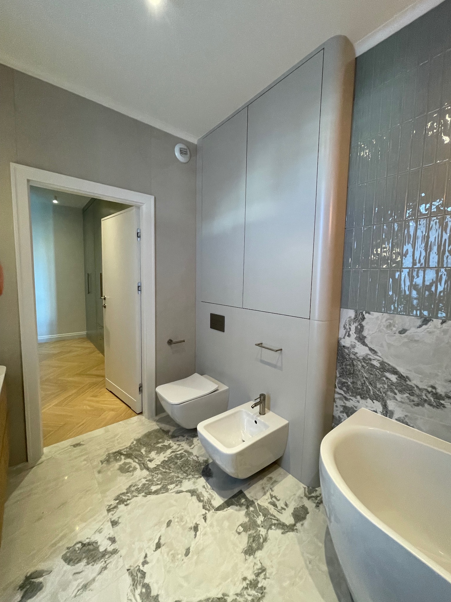 Nowoczesna łazienka w odcieniach szarości z marmurową podłogą i błyszczącymi, pionowymi płytkami, z białą armaturą: toaleta, bidet i wanna, oraz zabudową meblową z ukrytą spłuczką.
