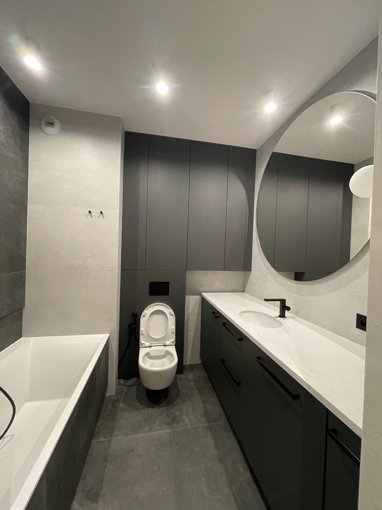 Nowoczesna łazienka w odcieniach szarości i bieli z owalnym lustrem, czarną armaturą i zabudową meblową pod sufit.
