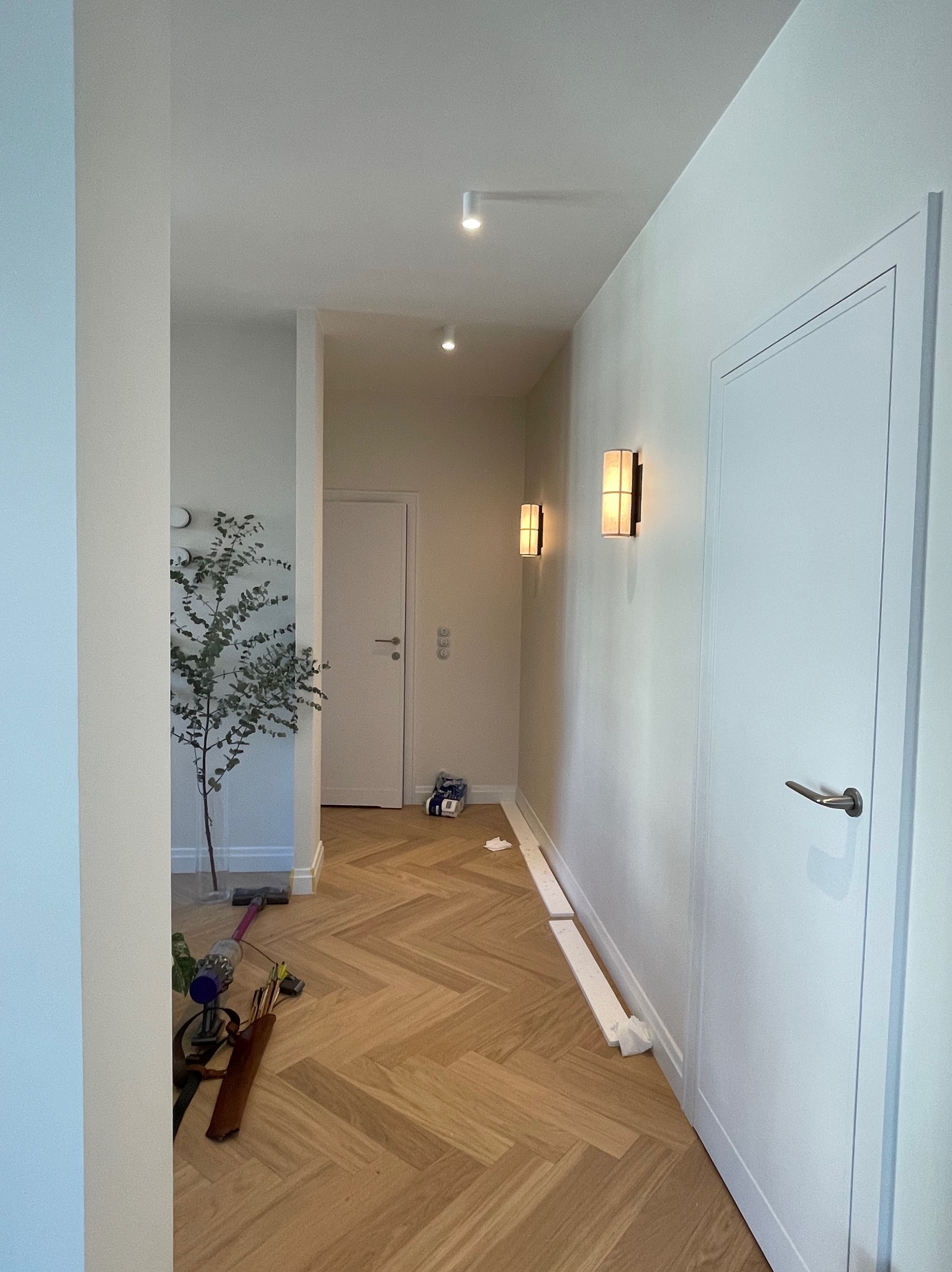 Wnętrze mieszkania w trakcie prac wykończeniowych: widoczna podłoga ułożona w jodełkę, białe ściany z zamontowanymi lampami ściennymi, drzwi wewnętrzne, narzędzia i materiały budowlane na podłodze,...