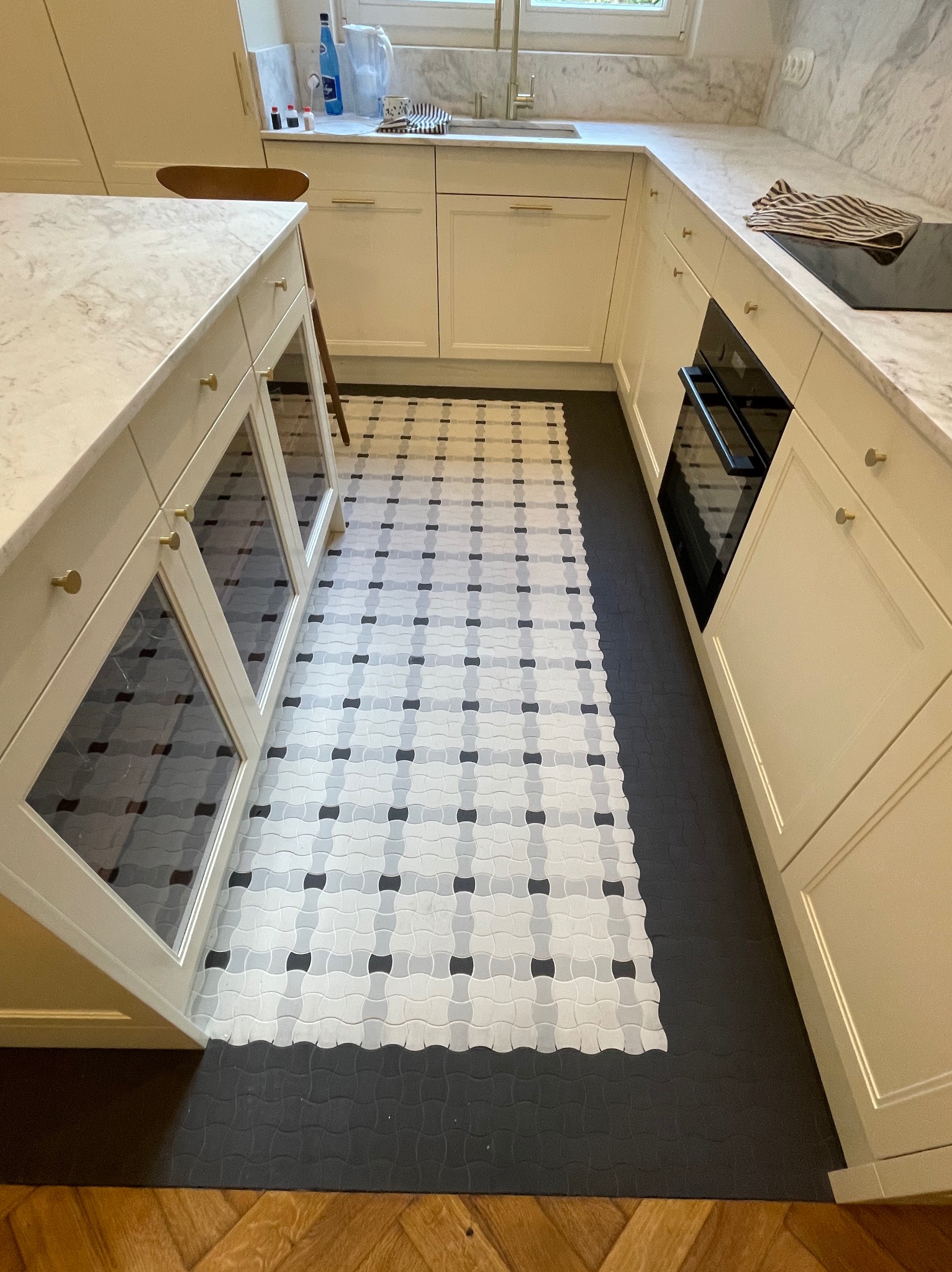 Wykończona kuchnia z kremowymi szafkami, marmurowym blatem i wzorzystą podłogą z płytek w odcieniach szarości i bieli, kontrastującą z ciemną, gumową podłogą graniczącą z parkietem.