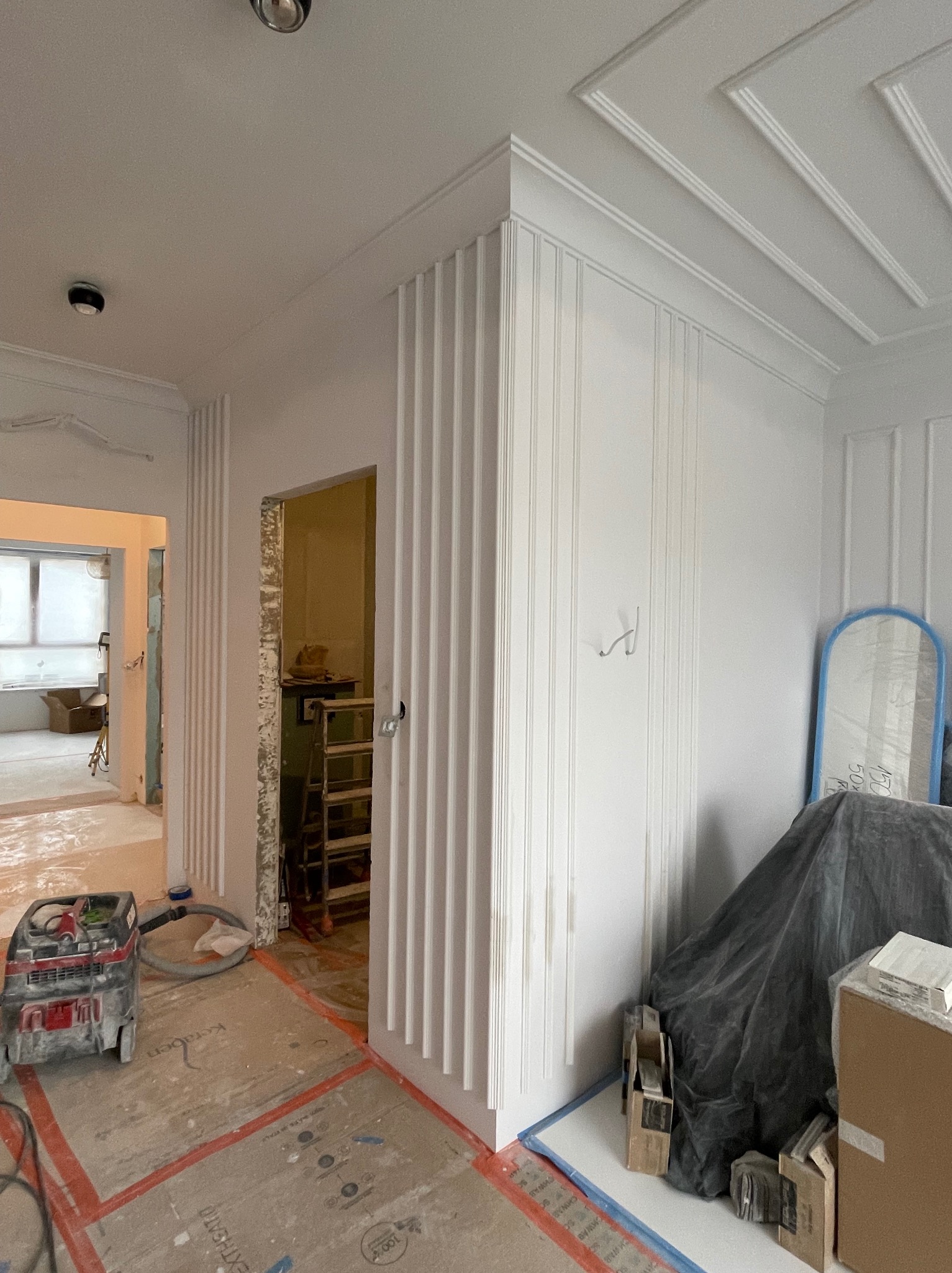 Wnętrze w trakcie prac wykończeniowych: ściany z dekoracyjnymi listwami pionowymi, widoczny odkurzacz przemysłowy, zabezpieczona podłoga, drabina w przejściu do innego pomieszczenia, elementy...