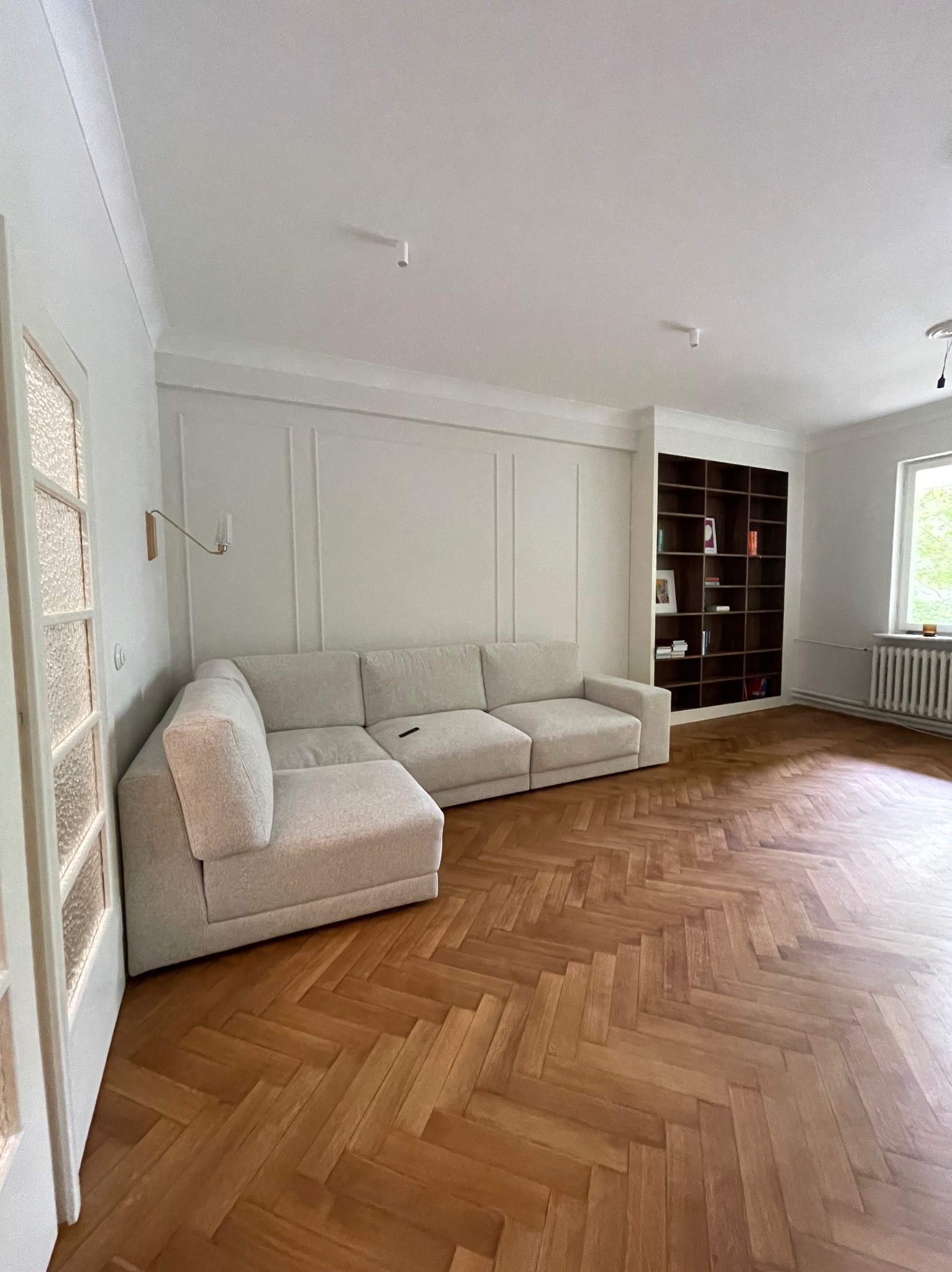 Jasny narożnik w przestronnym pokoju z parkietem w jodełkę, białe ściany z dekoracyjnymi listwami, ciemny regał z książkami w zabudowie.