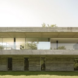 Swisstec Okna i Drzwi - Dwukondygnacyjny dom z betonu architektonicznego, z dużymi oknami i przeszkloną balustradą balkonu na piętrze, otoczony trawnikiem.