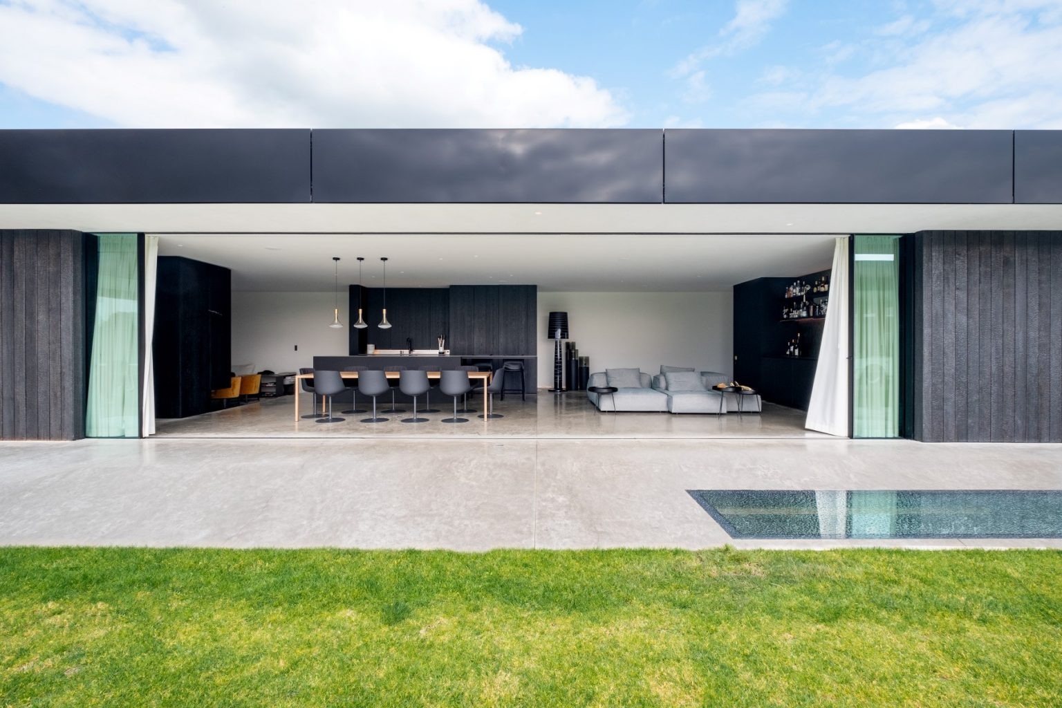 Nowoczesny dom z dużymi, przesuwnymi drzwiami tarasowymi, otwierającymi przestrzeń na zewnątrz, widok na minimalistyczne wnętrze z kuchnią i salonem, trawnik w pierwszym planie.