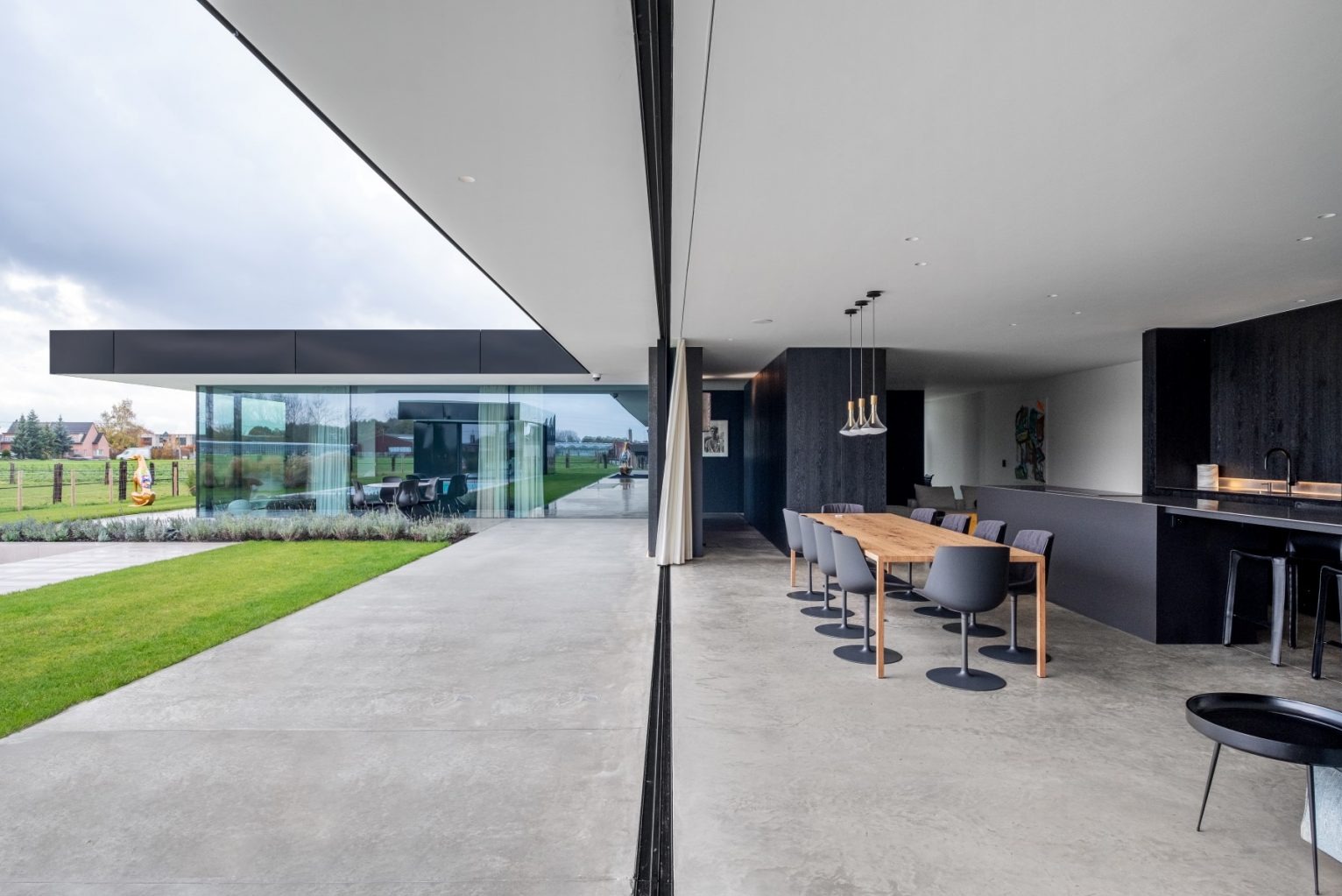 Nowoczesny dom z panoramicznymi przesuwnymi drzwiami, widok na ogród, minimalistyczne wnętrze z drewnianym stołem i czarną zabudową kuchenną.