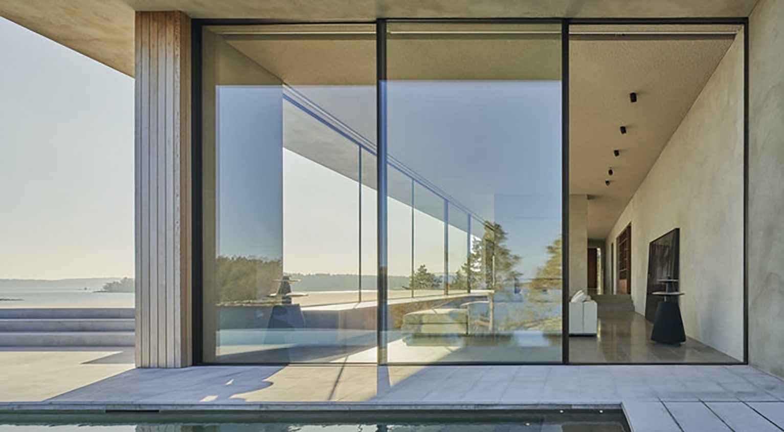 Panoramiczne, trzyskrzydłowe drzwi przesuwne z czarnymi ramami, otwierające się na taras z basenem i widokiem na jezioro, w minimalistycznym domu z betonowymi ścianami i drewnianymi elementami.